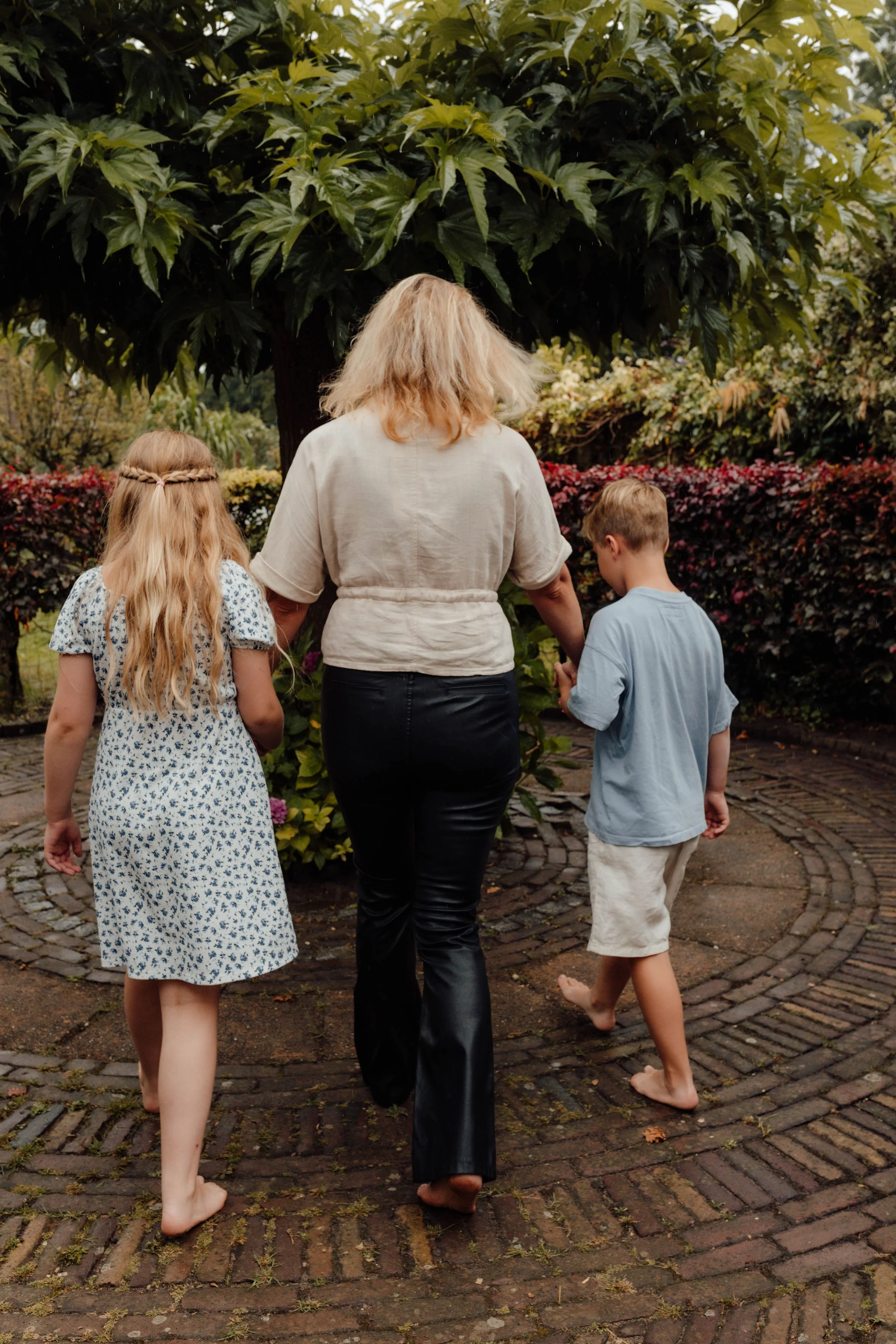 Drie mensen, een bonusmoeder en twee bonuskinderen, lopen hand in hand op een geplaveide paden in een tuin of park, omgeven door groene planten en bloemen.