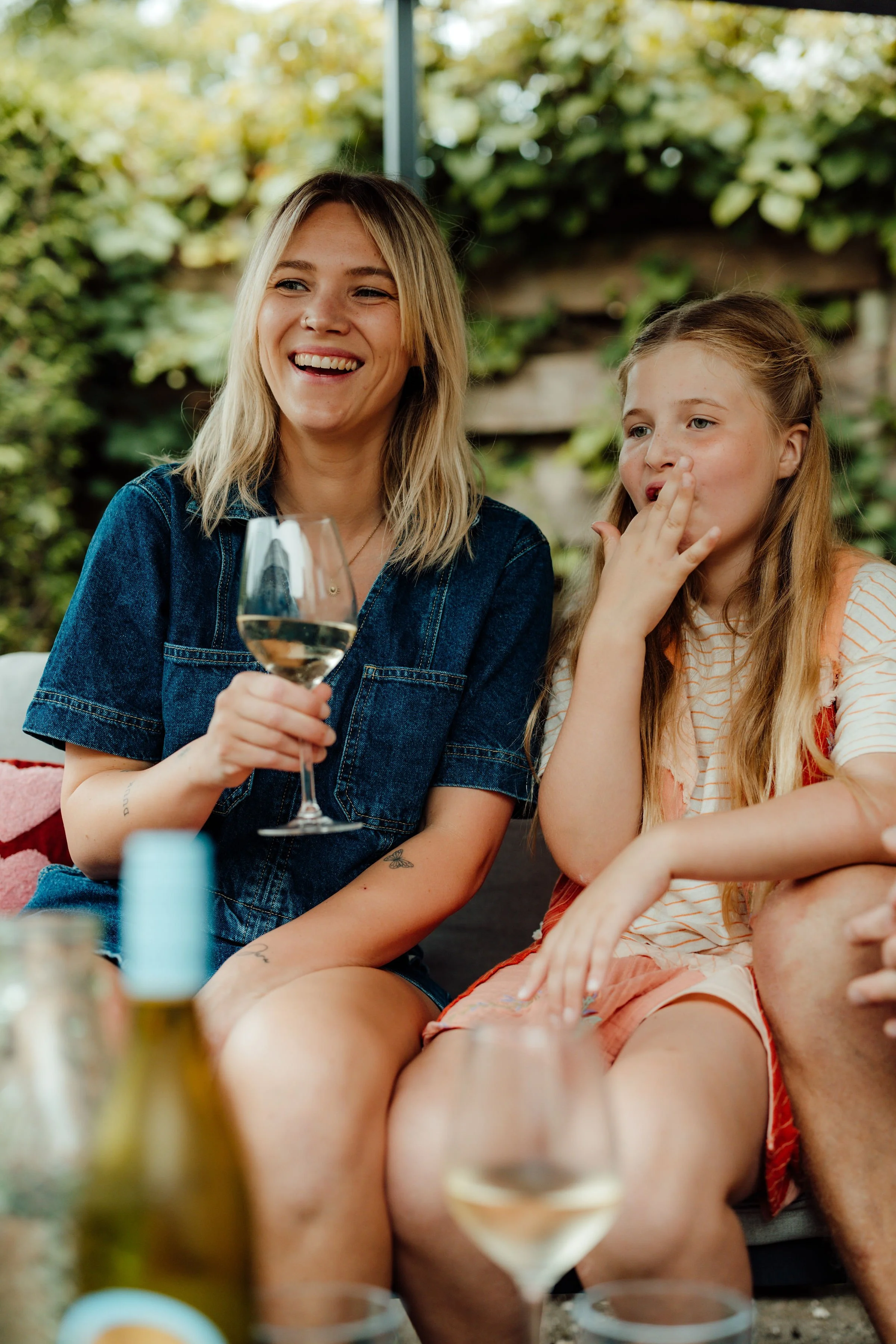 Moeder en dochter genieten samen buiten. Eén vrouw houdt een glas witte wijn vast en lacht, de andere jongen is aan het hapje en de tweede vrouw is niet zichtbaar.