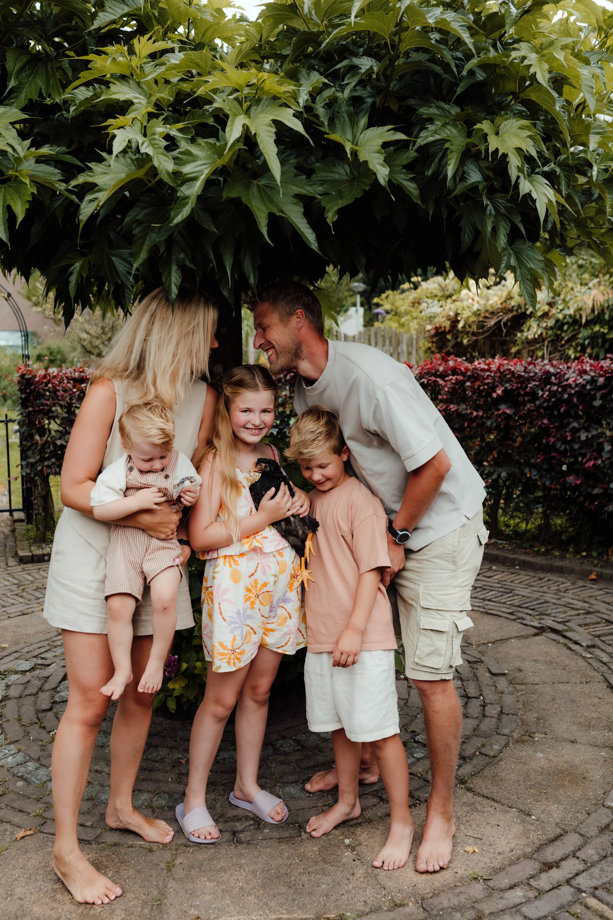 Samengesteld gezin met vier kinderen staat buiten onder een grote boom, ze lachen en knuffelen met een zwarte kip in hun midden.