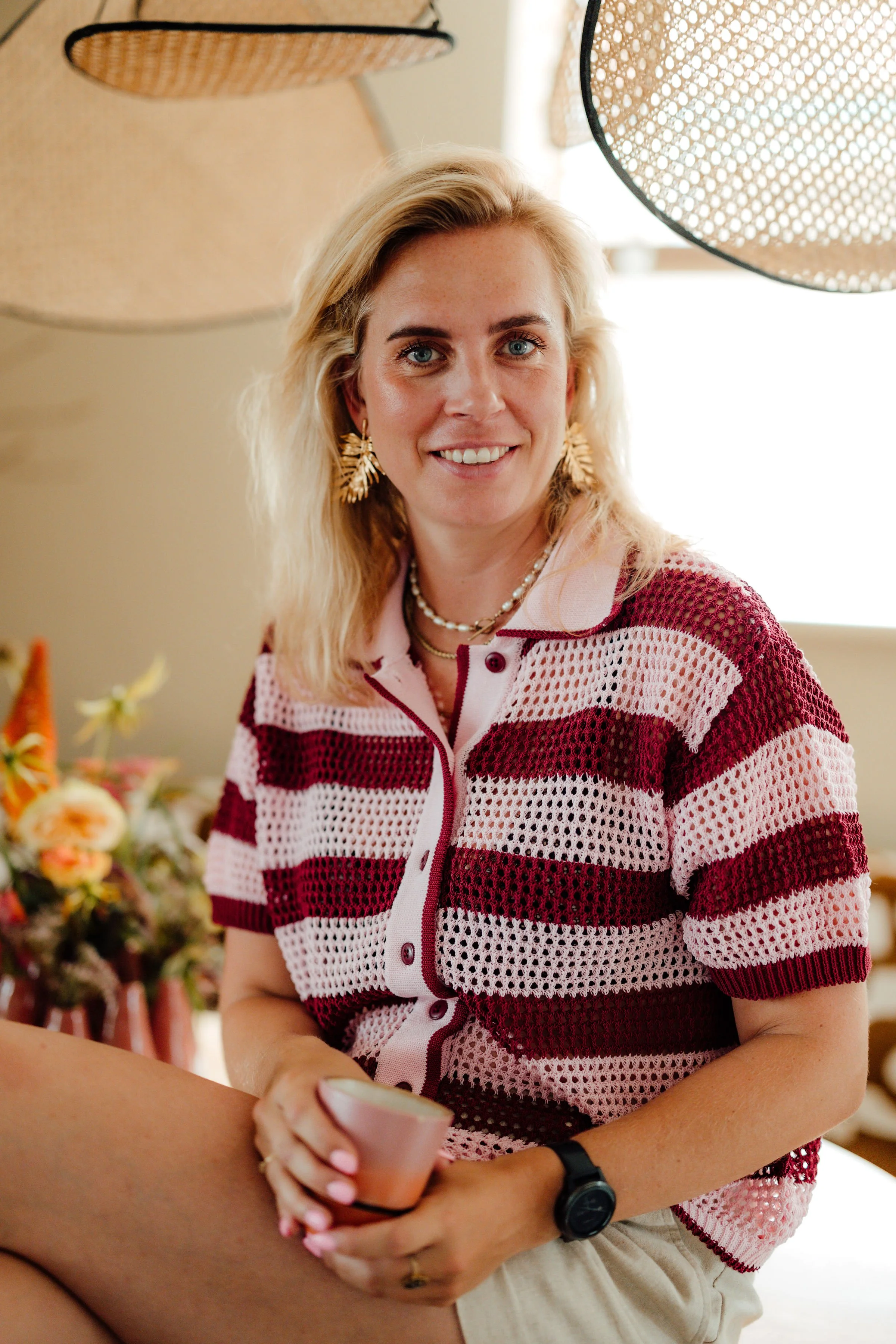 Een glimlachende blonde bonusmoeder in een roze en maroon gebreide blouse met sieraden en een horloge, zittend binnenshuis met zachte verlichting.