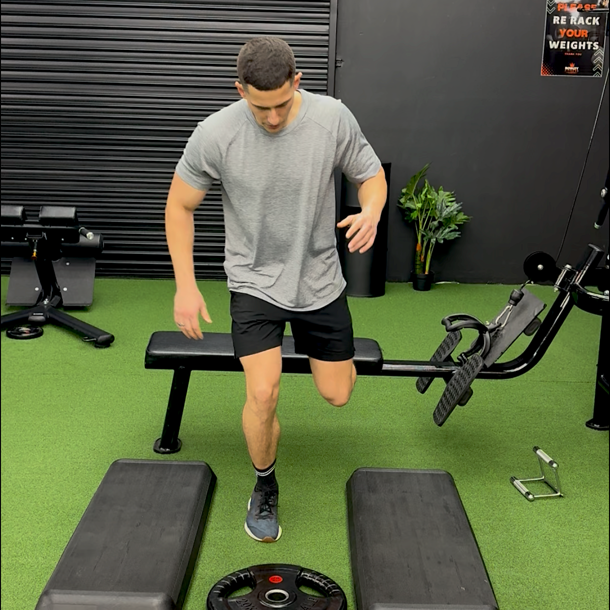 A man performing a step-up exercise in a gym, stepping onto a raised platform with one leg. The gym has green turf flooring, workout equipment, and a motivational poster on the wall.
