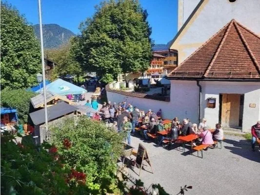 Menschen sitzen an Tischen im Freien vor einer weißen Kirche mit rotem Ziegeldach, umgeben von Bäumen, bei sonnigem Wetter.