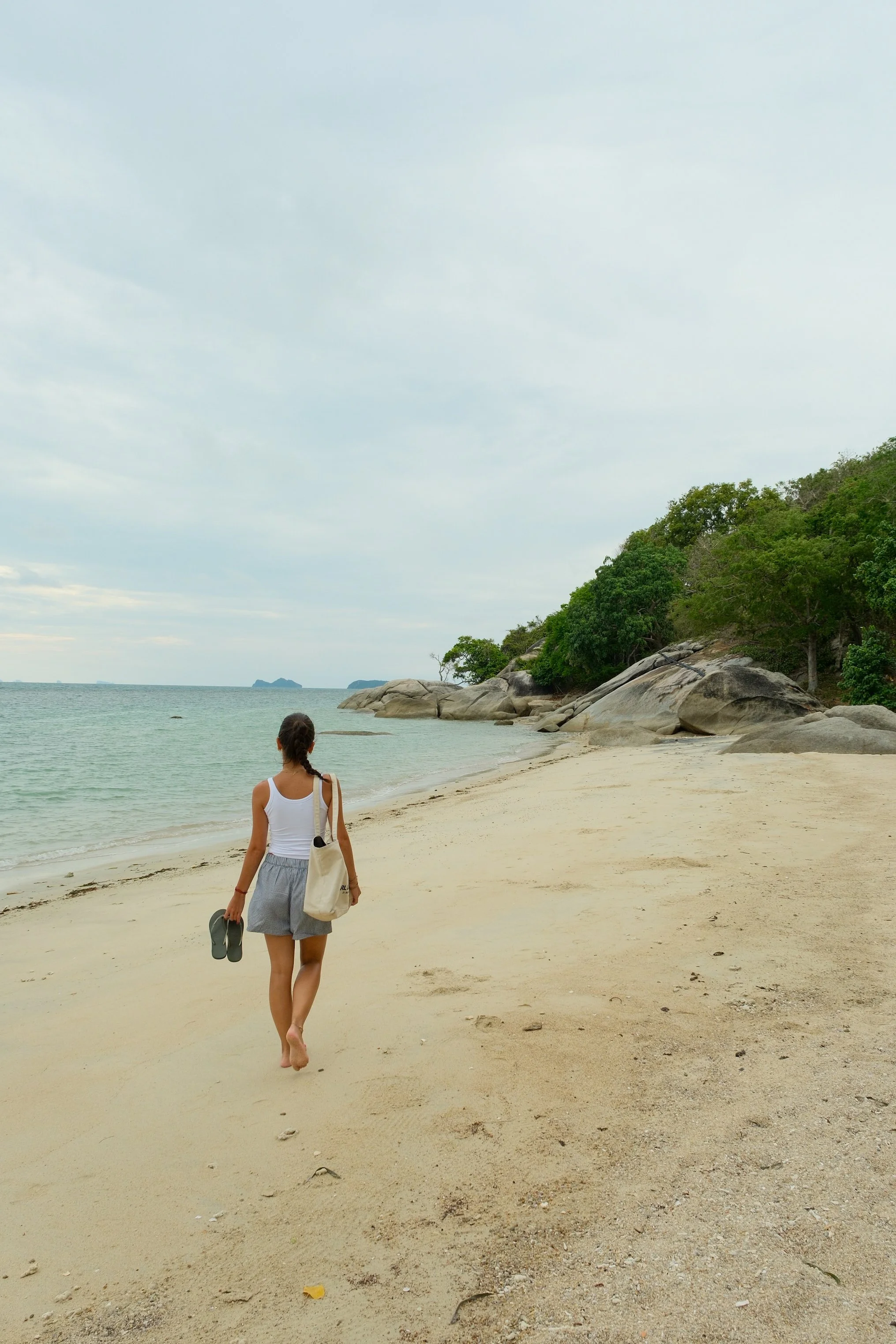Guest walking barefoot along a secluded sandy beach framed by calm waters, tropical forest, and granite rocks