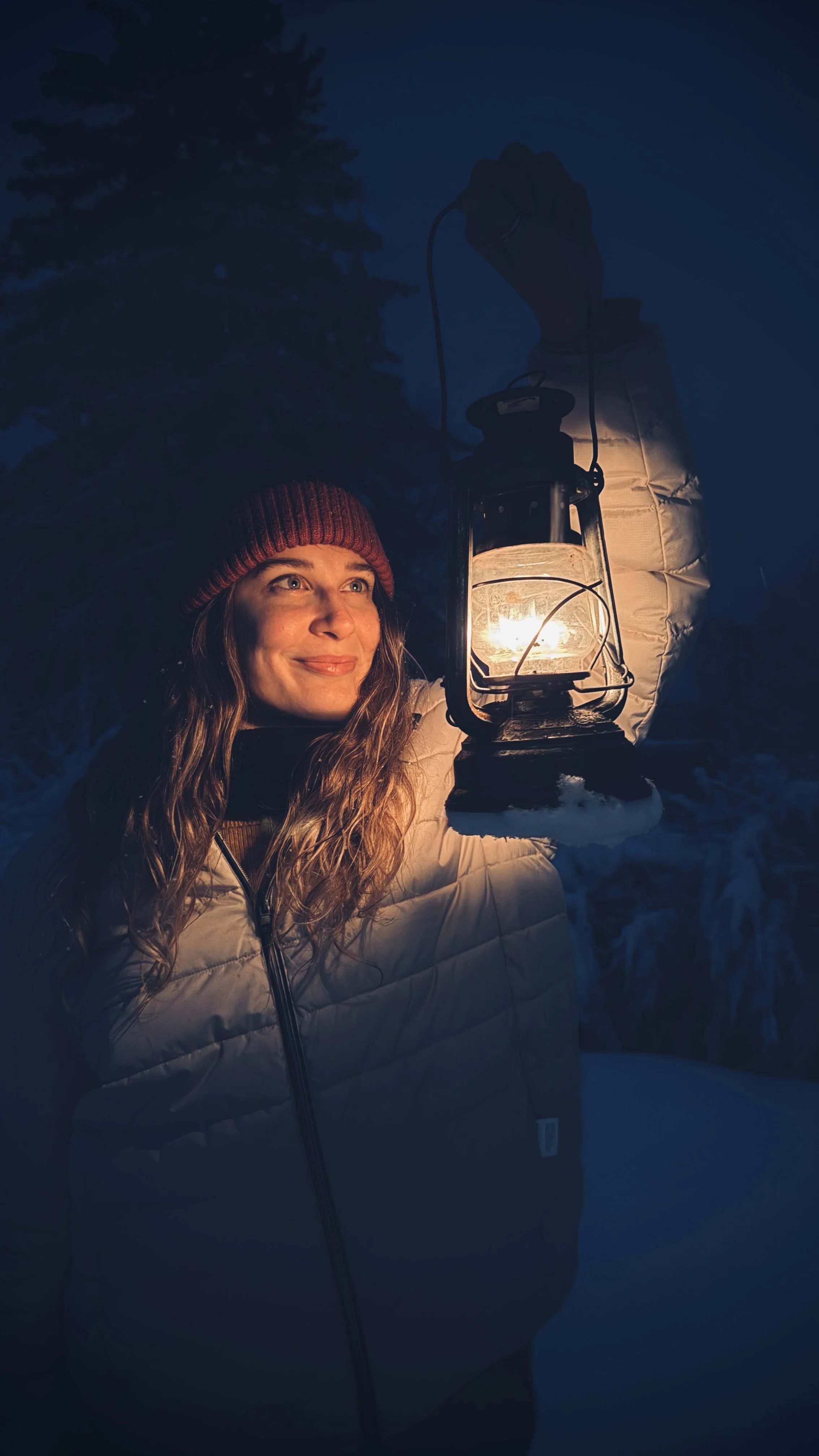 A guest holding a lantern at dusk, standing in fresh snow as the forest fades into blue evening light, Nutchel Forest Village Alsace – Plaine, France.