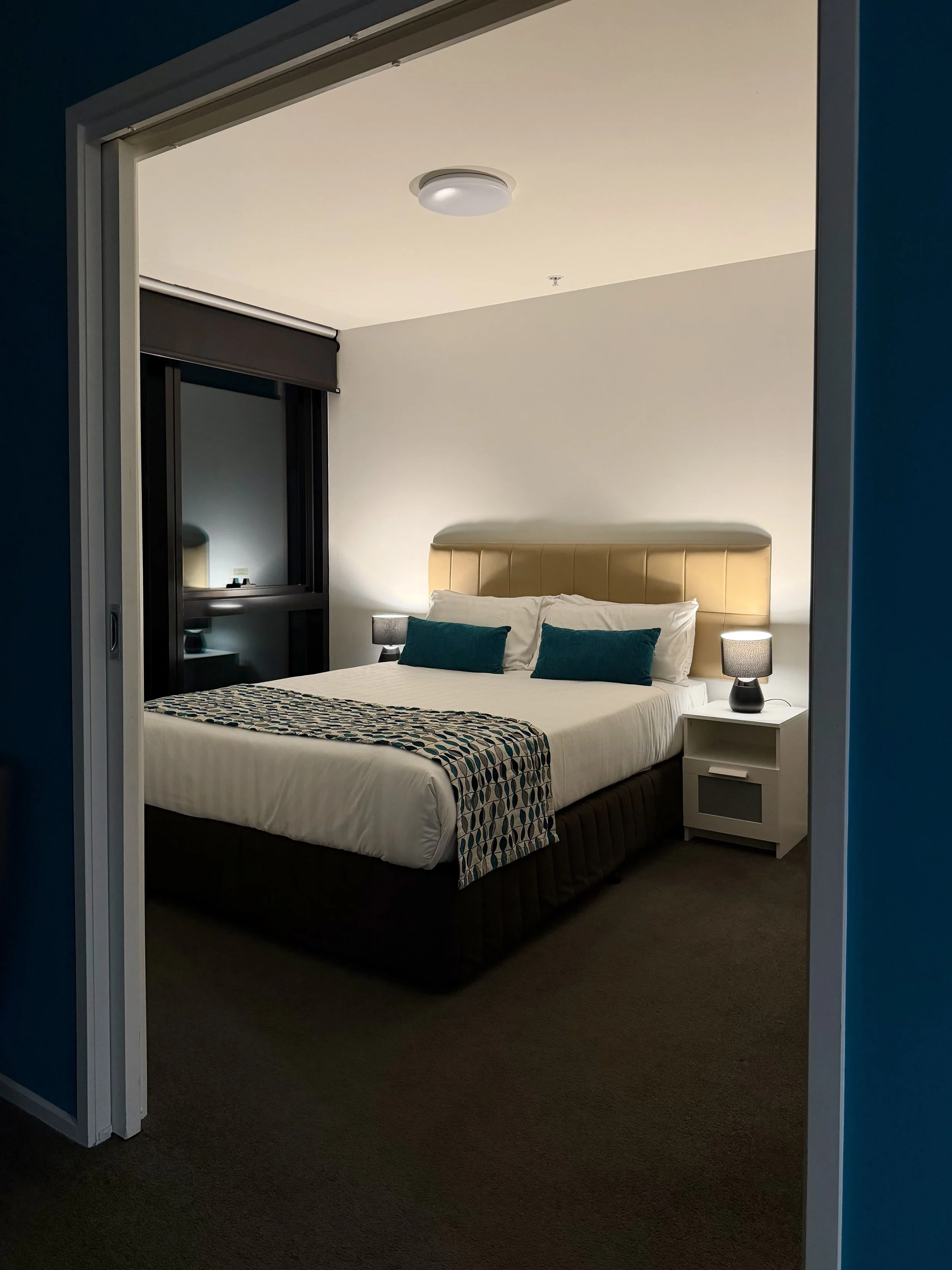 Modern hotel bedroom featuring a king-size bed, beige upholstered headboard, teal cushions, and warm bedside lighting
