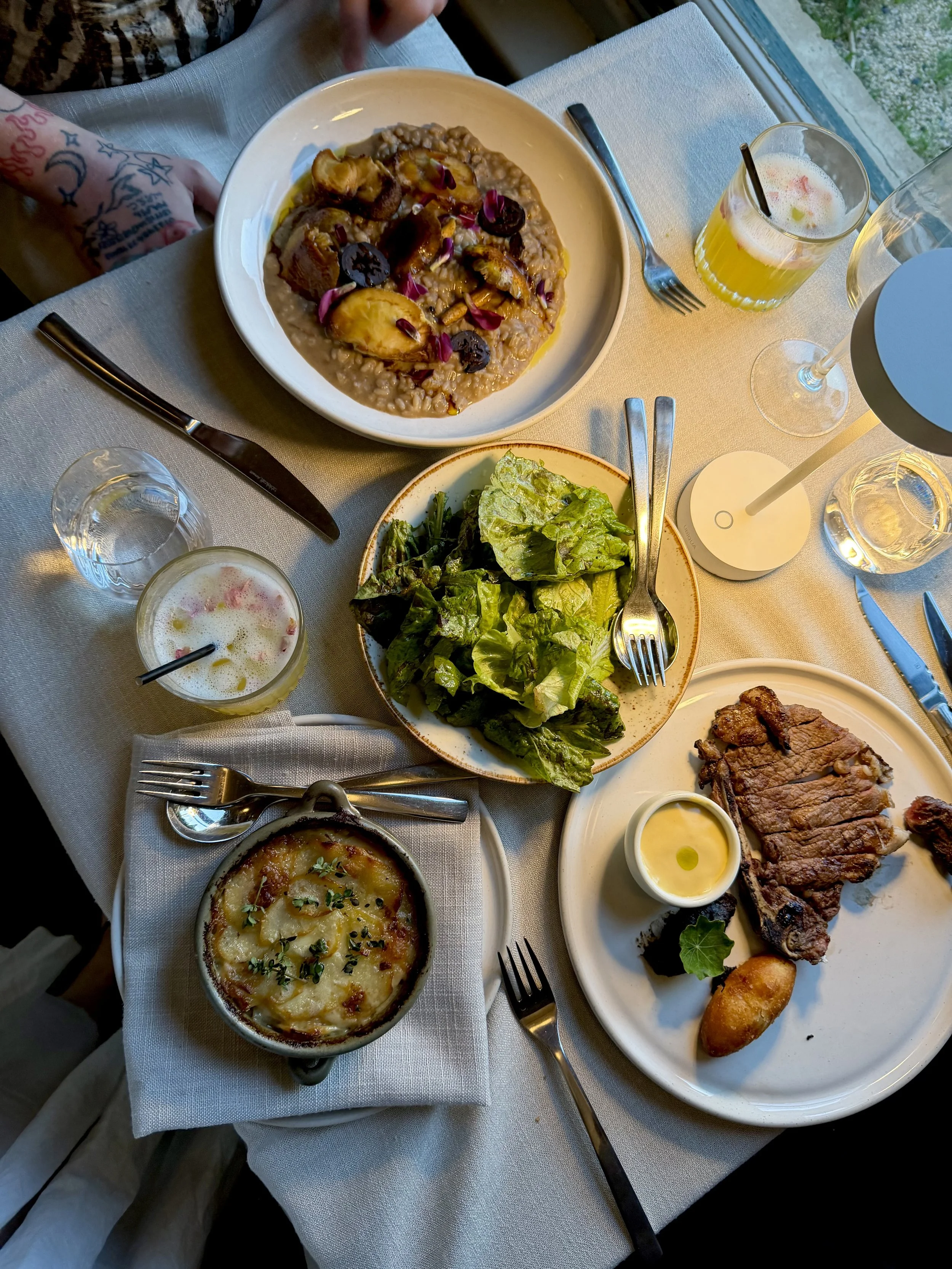 Restaurant dining table featuring grilled steak, fresh salad, creamy potato gratin, and refreshing drinks