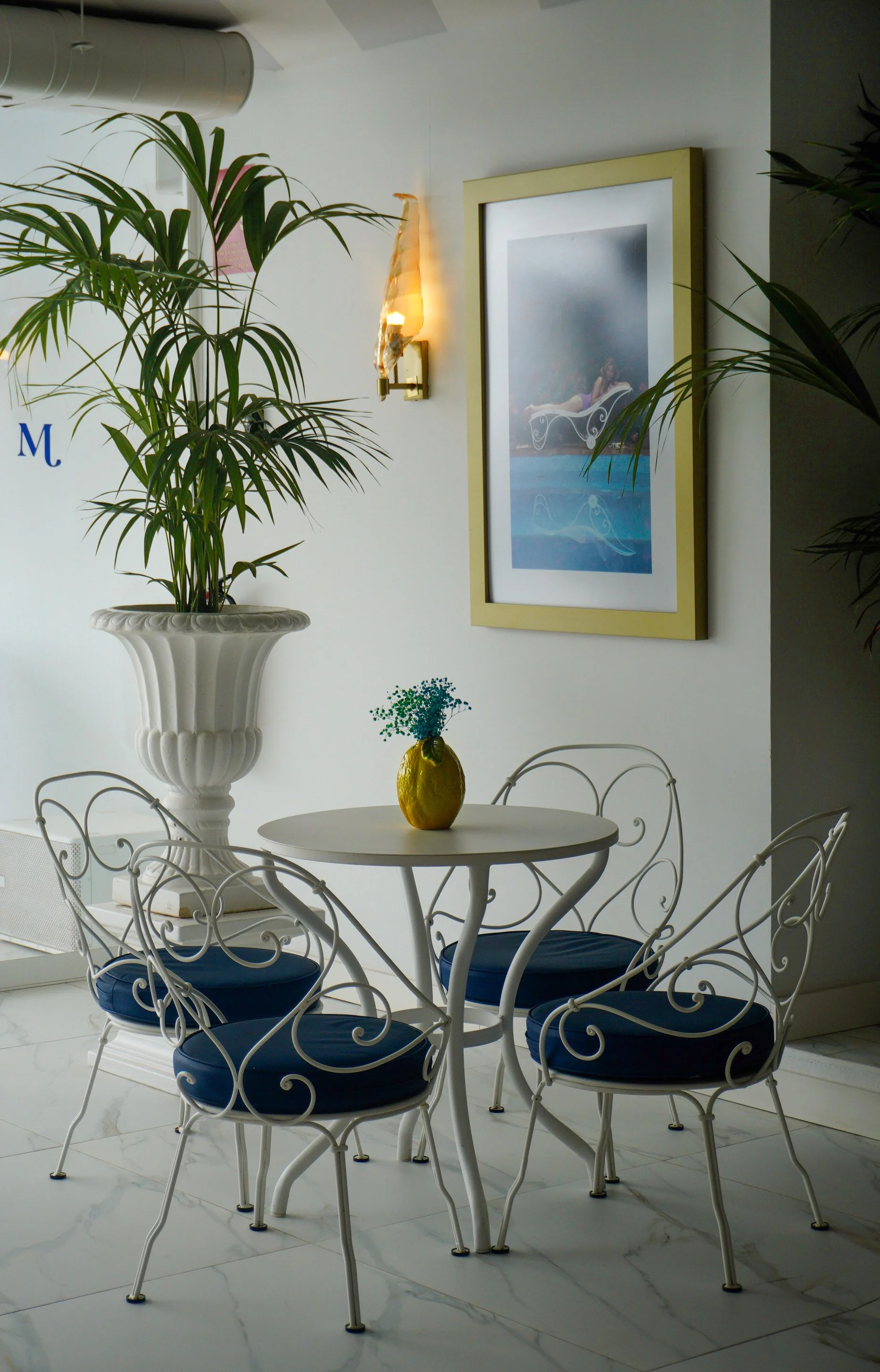Elegant lounge area with white wrought iron furniture, blue cushions, tropical plants, and framed artwork