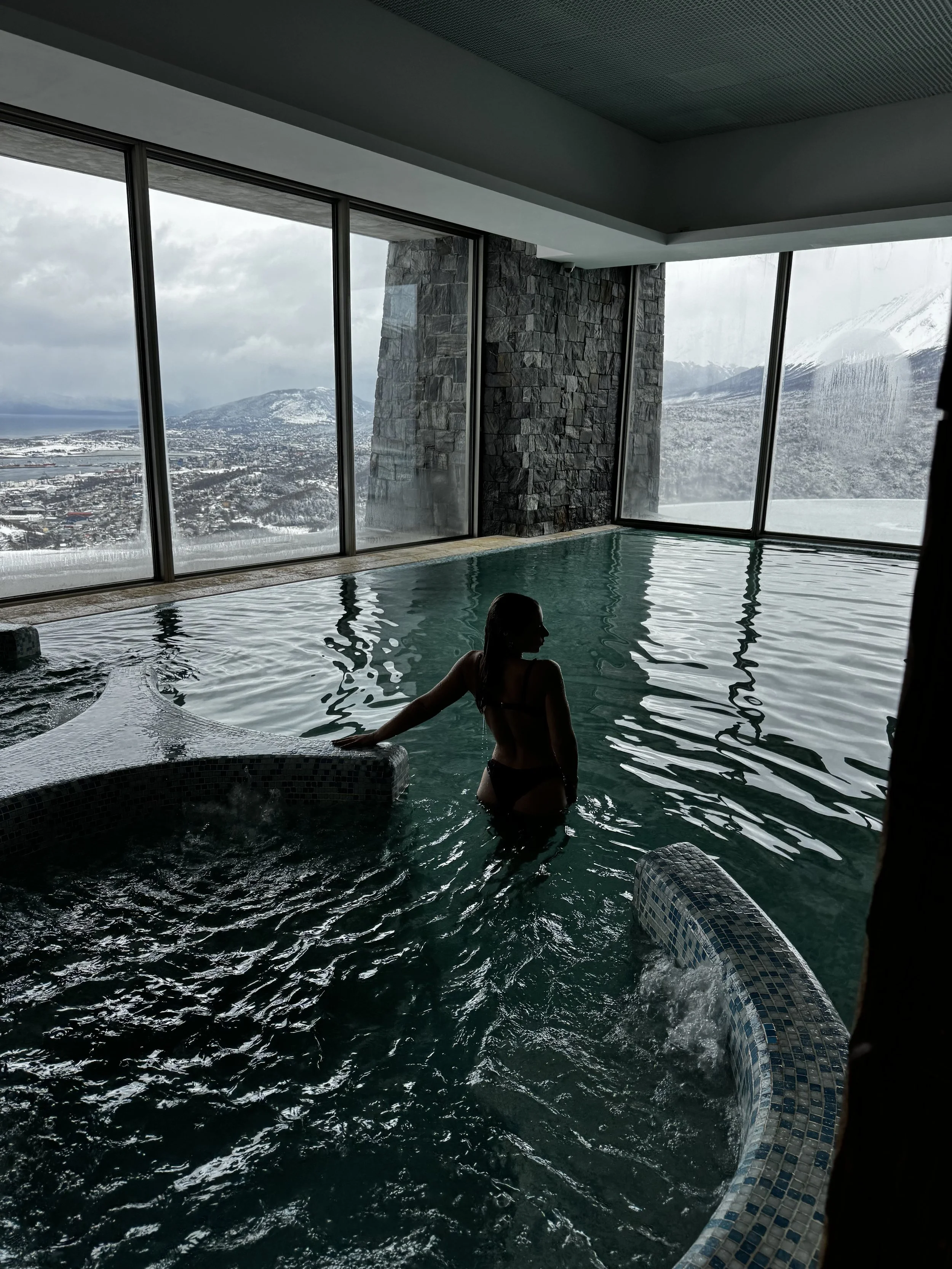 Guest relaxing in an indoor pool with panoramic views across snowy mountains, Ushuaia, and the Beagle Channel