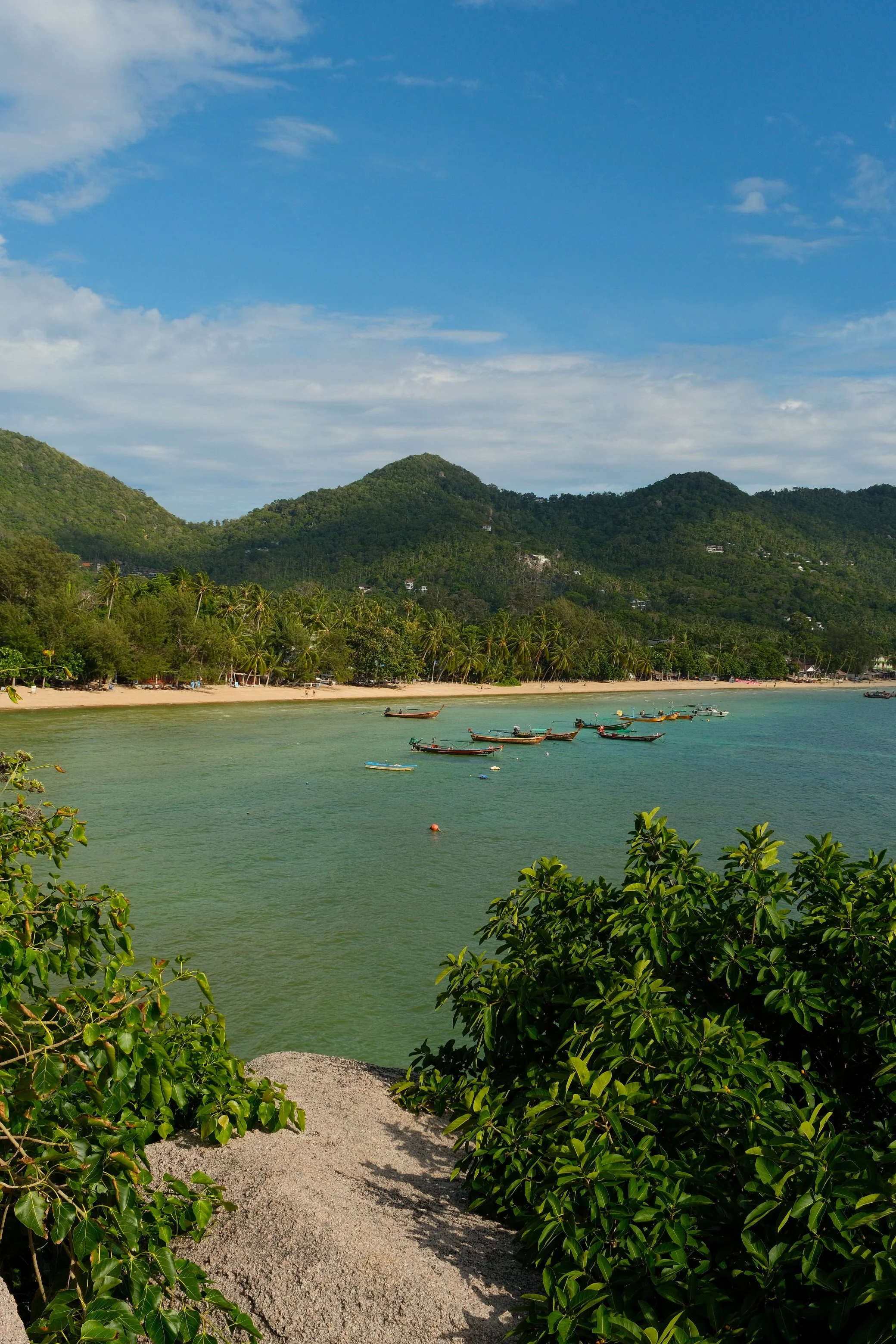 Elevated view across a tropical bay with longtail boats, palm-lined shores, and forested hills