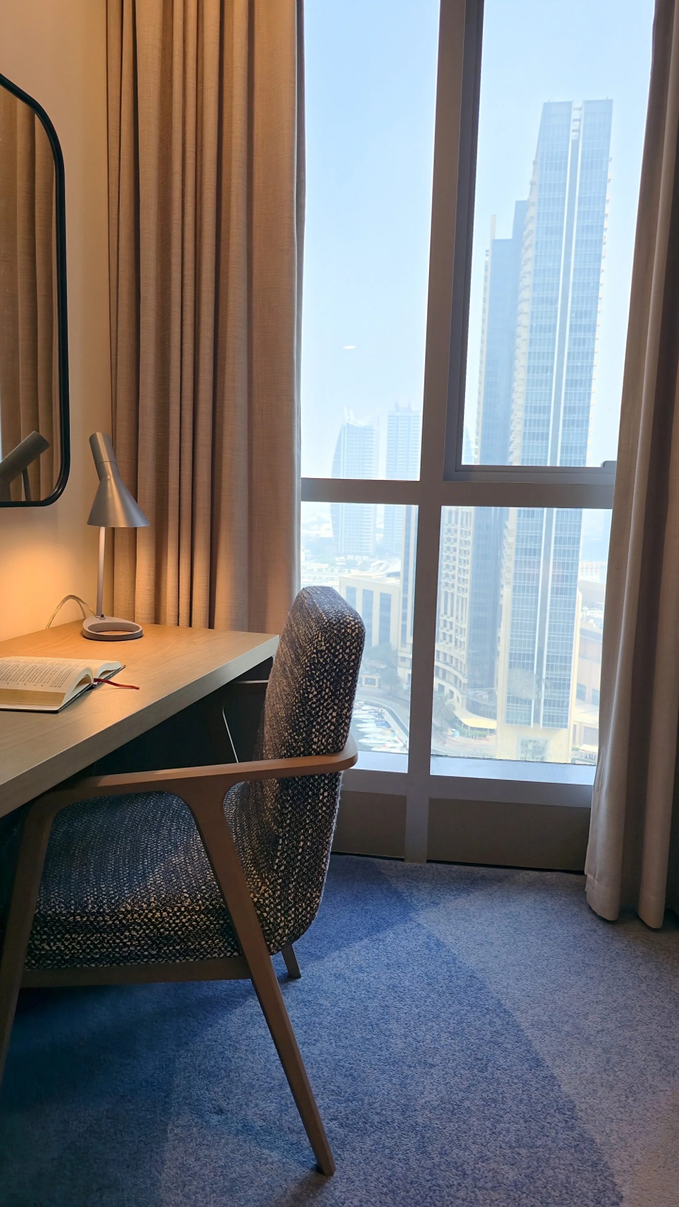 Elegant hotel room workspace featuring a wooden desk, upholstered chair, reading lamp, and floor-to-ceiling city views
