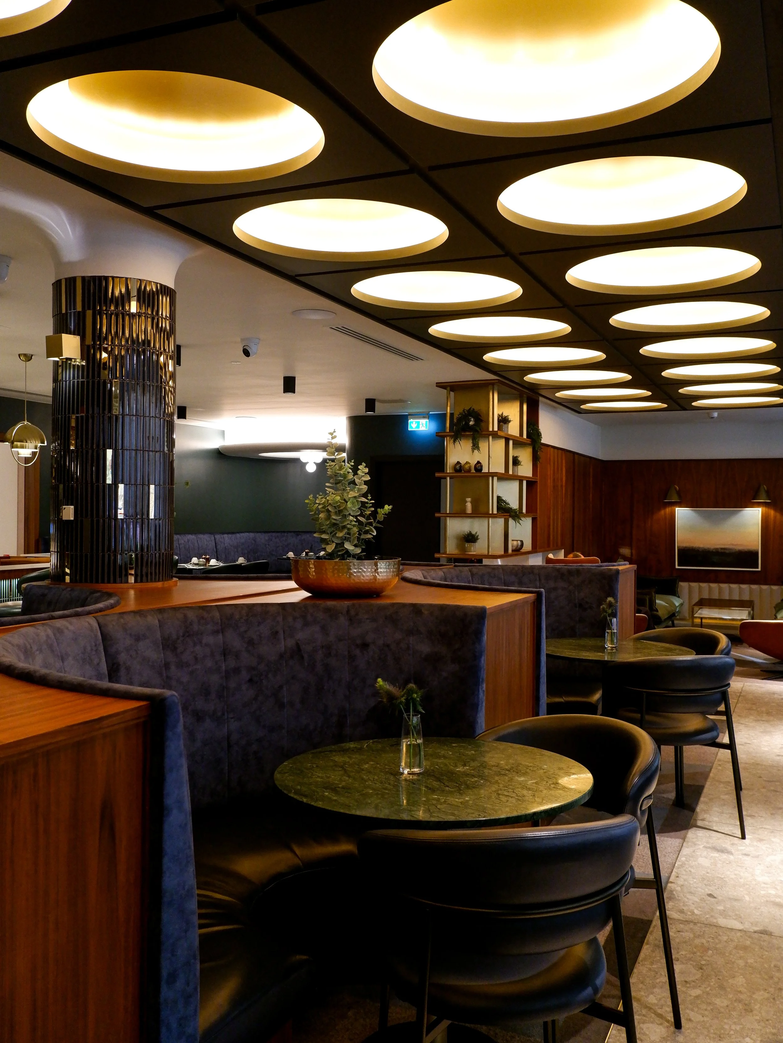 Elegant hotel restaurant featuring velvet booths, marble tables, dark wood finishes, and circular ceiling lights
