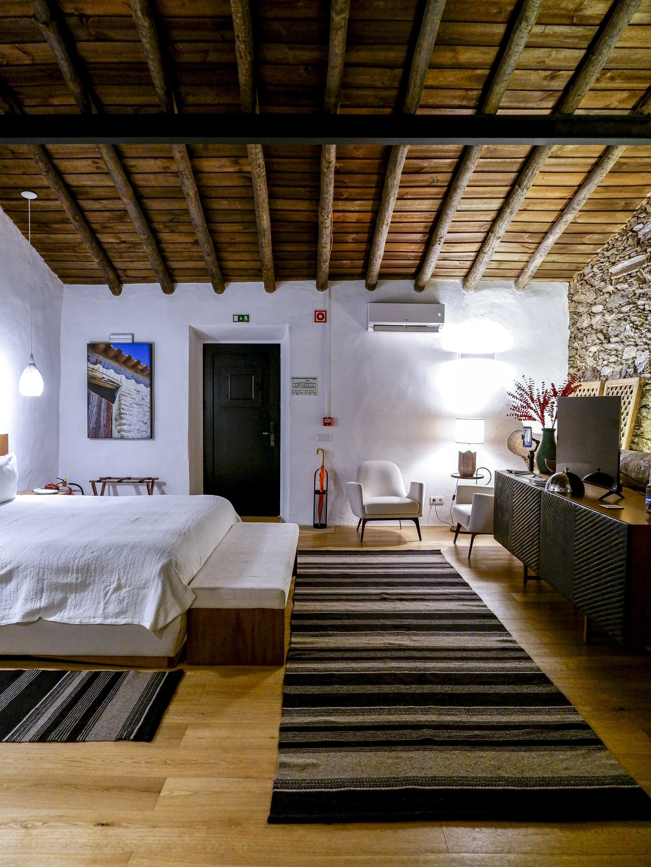 Rustic guest room with exposed wooden ceiling beams, stone wall accents, and warm minimalist furnishings creating a serene countryside atmosphere, Land of Alandroal – Alandroal, Portugal.