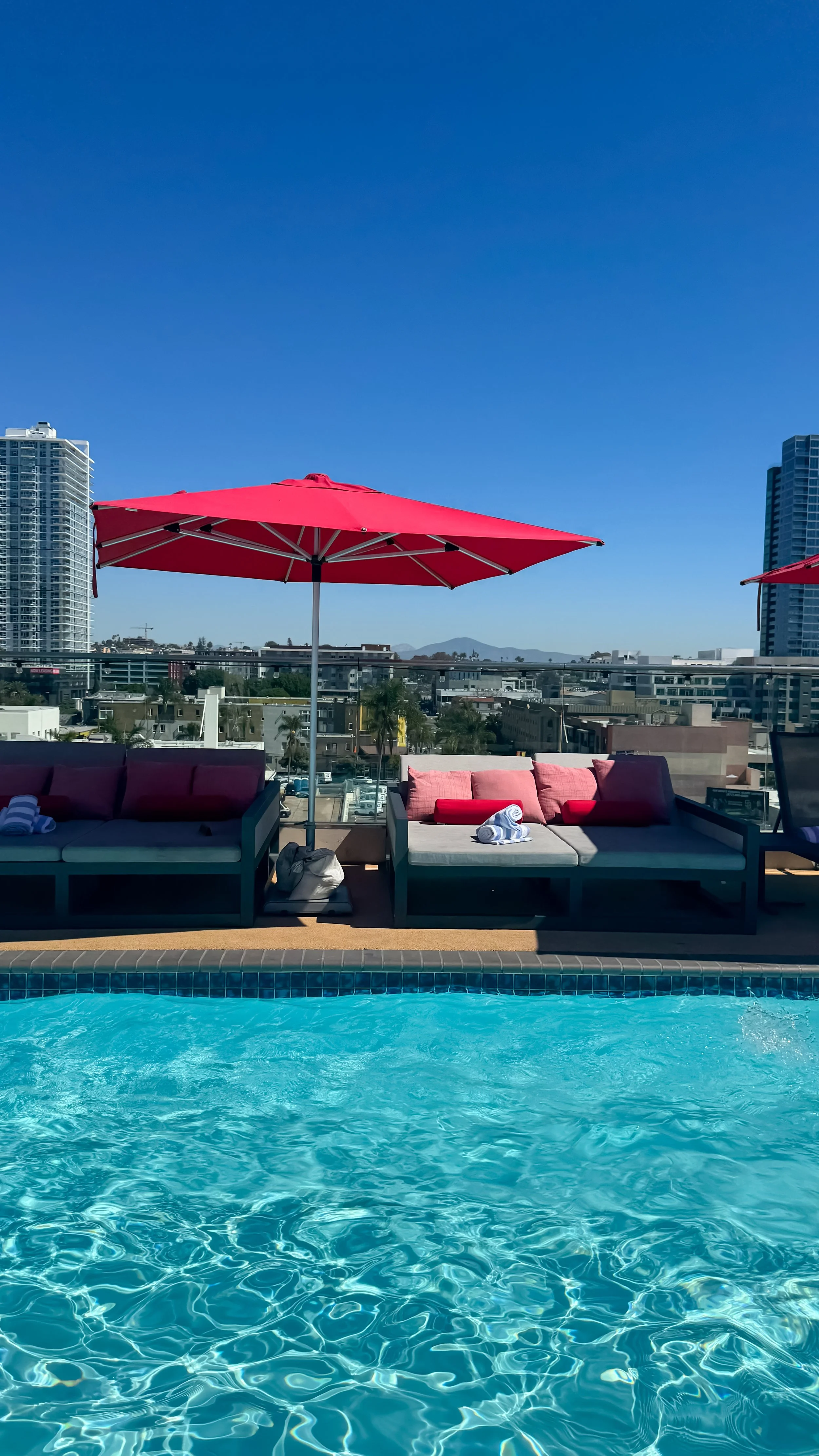 Sunny rooftop pool surrounded by red umbrellas, lounge beds, and sweeping city skyline views