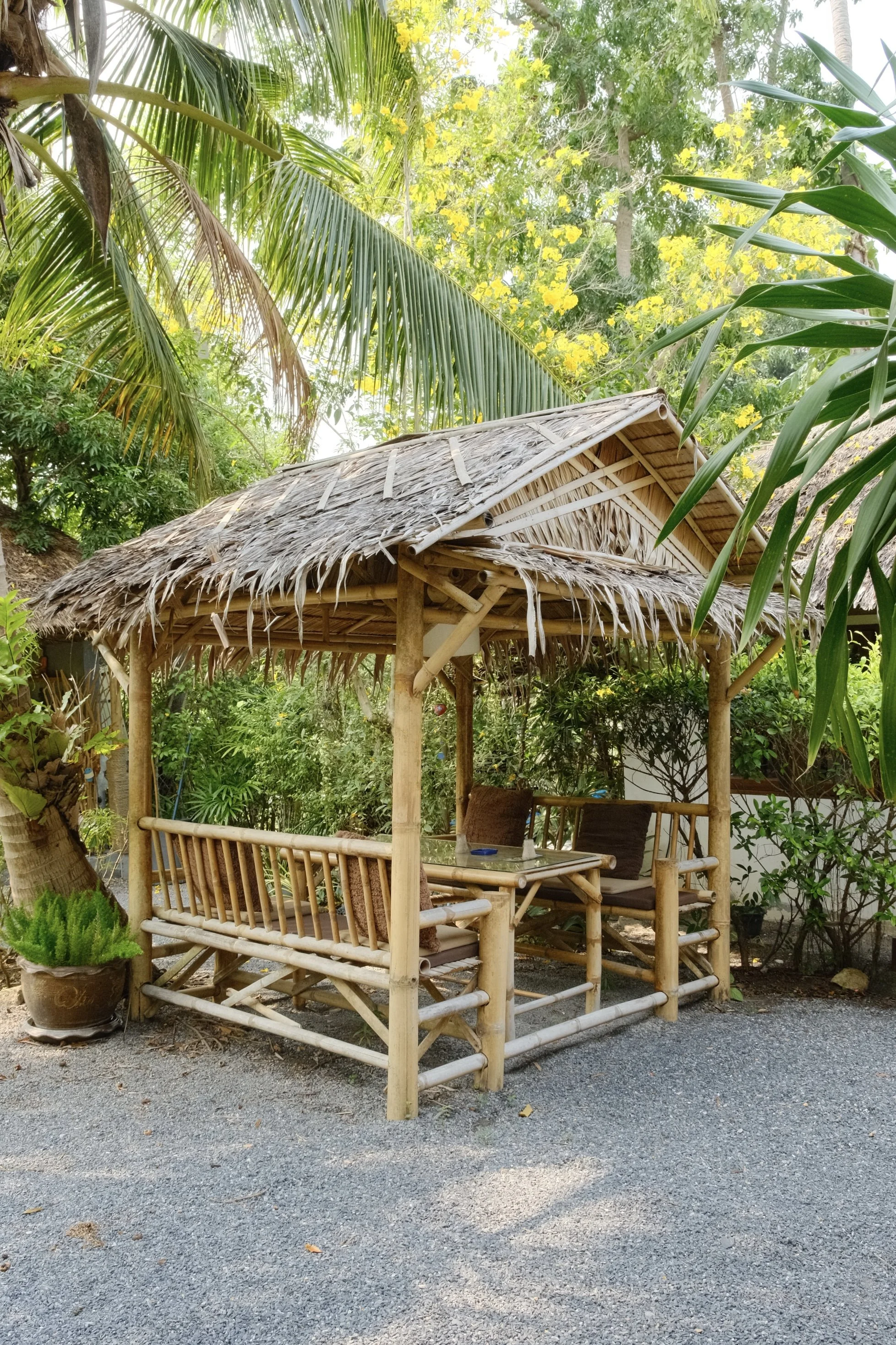 Traditional bamboo garden pavilion with cushioned seating beneath a thatched roof and tropical trees