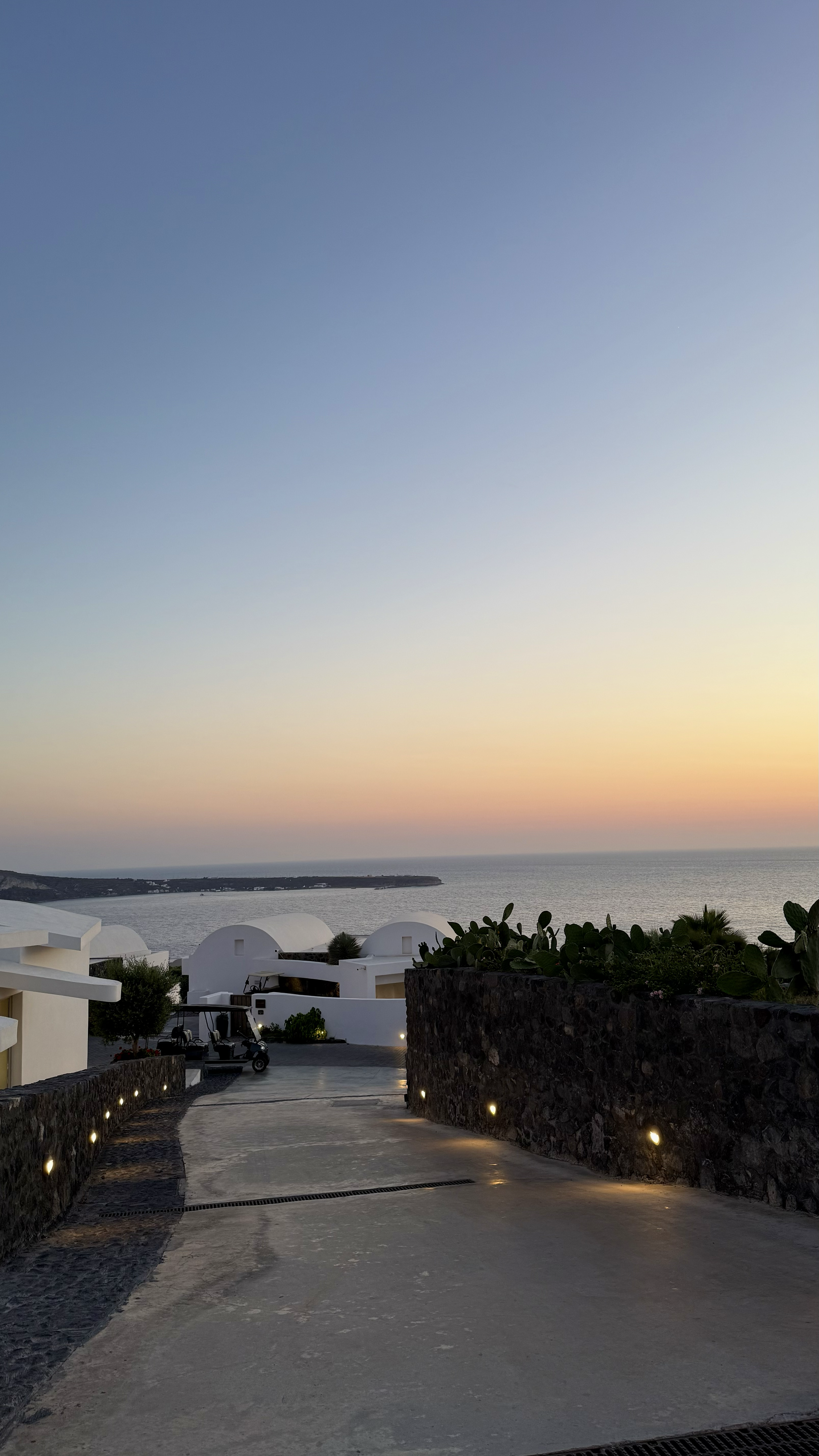 Peaceful resort pathway framed by volcanic stone walls, Cycladic architecture, and panoramic coastal scenery