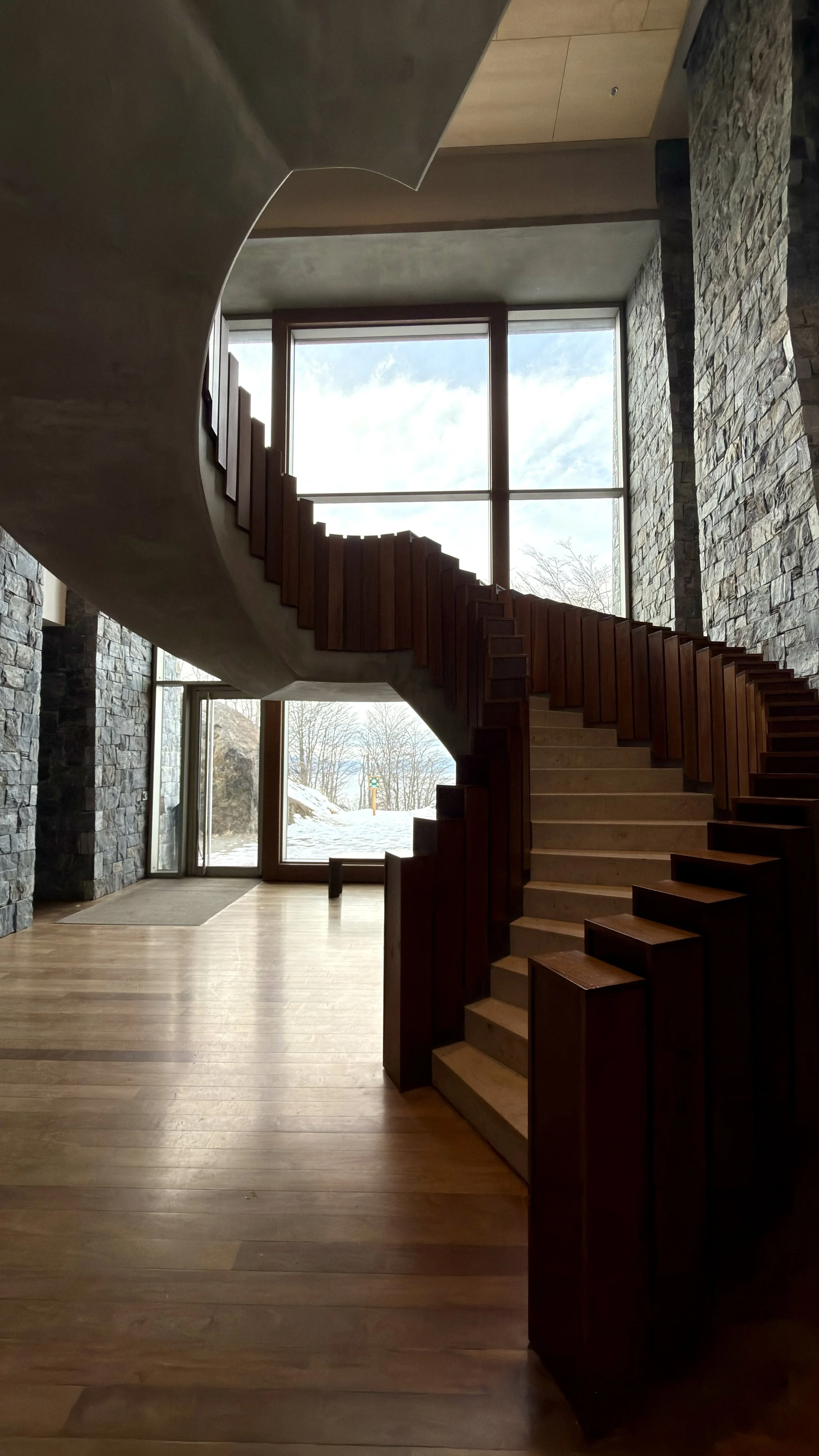 Sculptural curved staircase framed by natural stone walls, warm wood details, and floor-to-ceiling windows