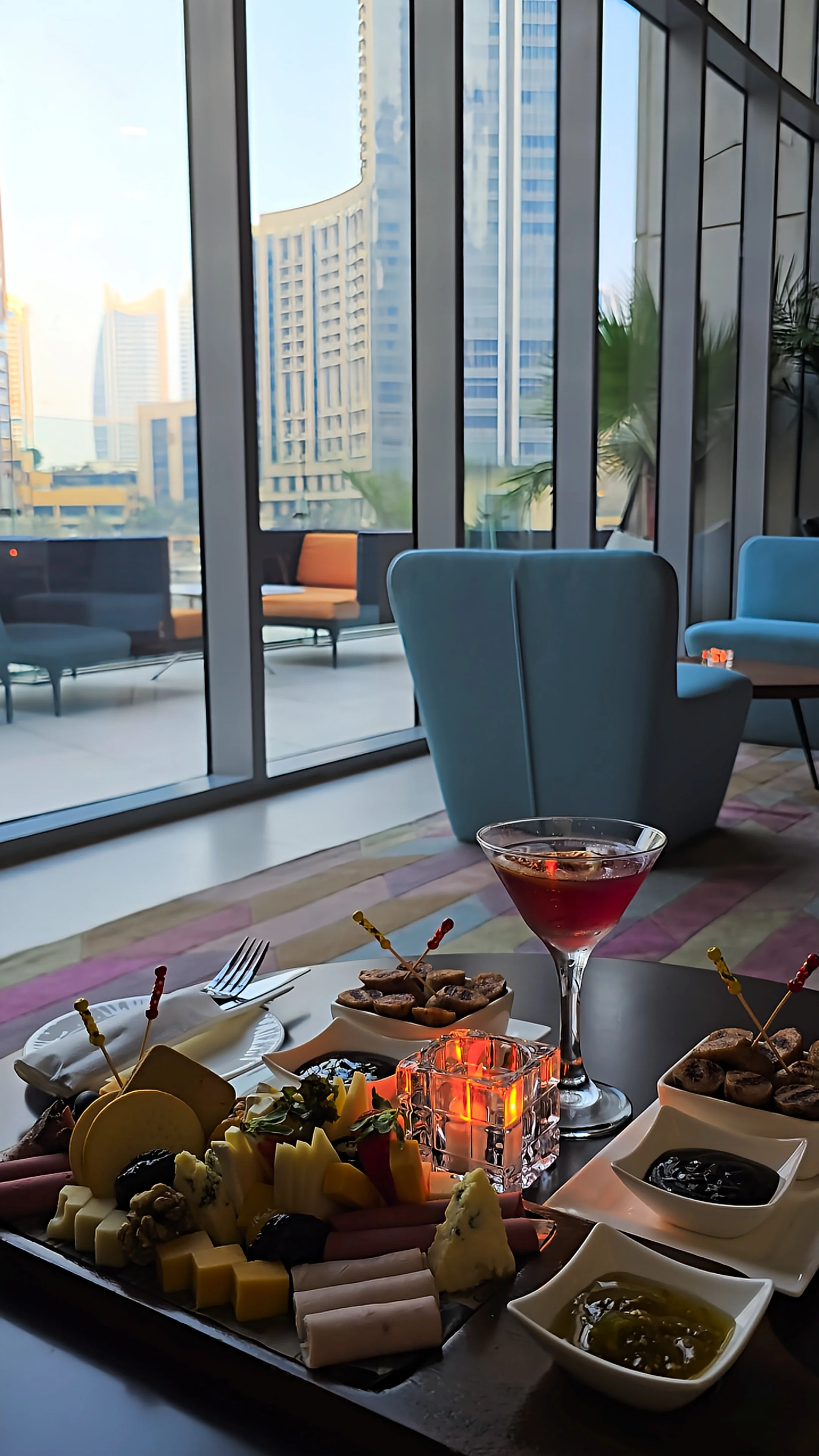 Elegant hotel lounge featuring a cheese and charcuterie platter, cocktail, candlelight, and floor-to-ceiling windows