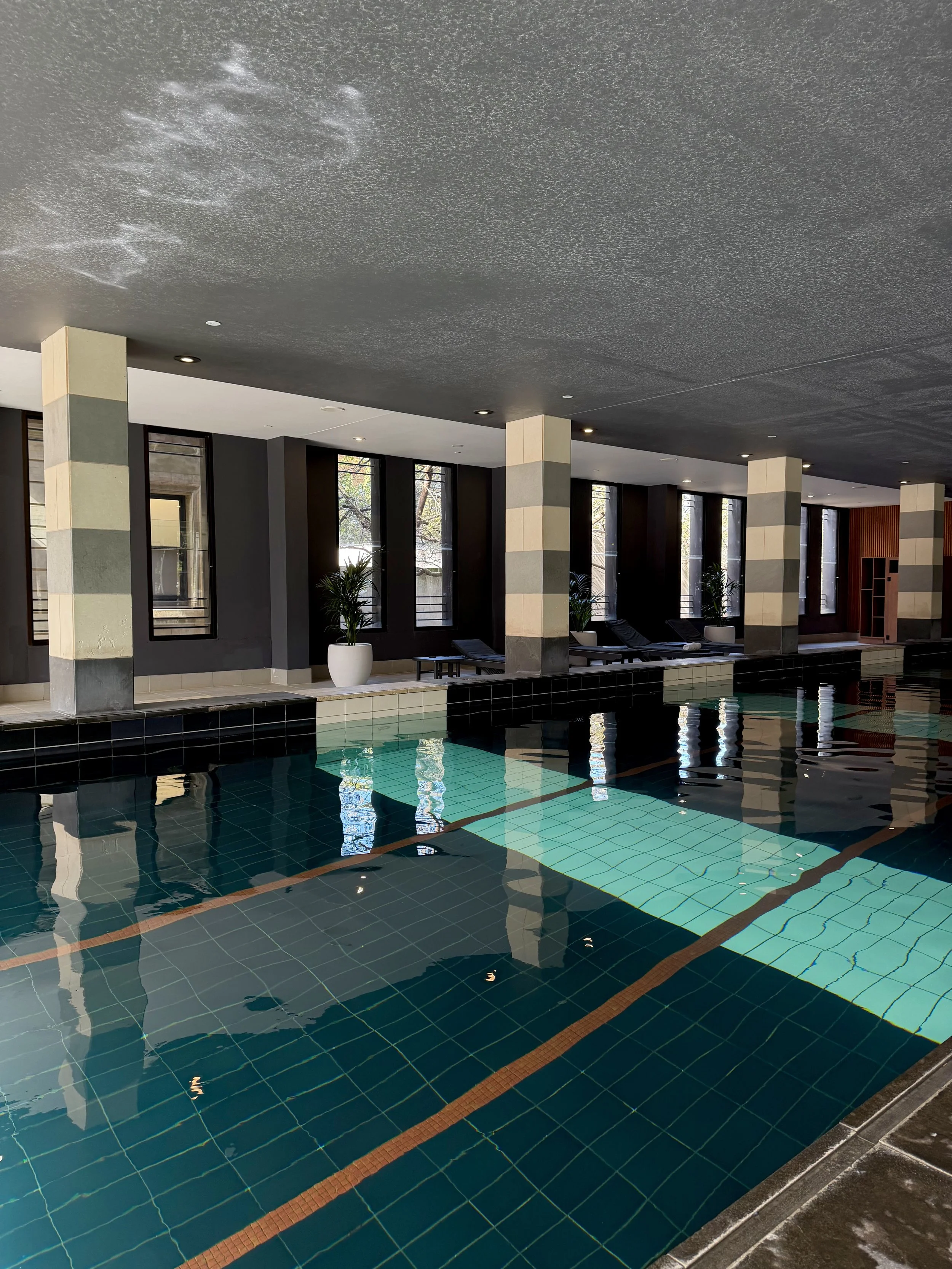 Indoor swimming pool featuring turquoise water, tiled columns, comfortable loungers, and natural light