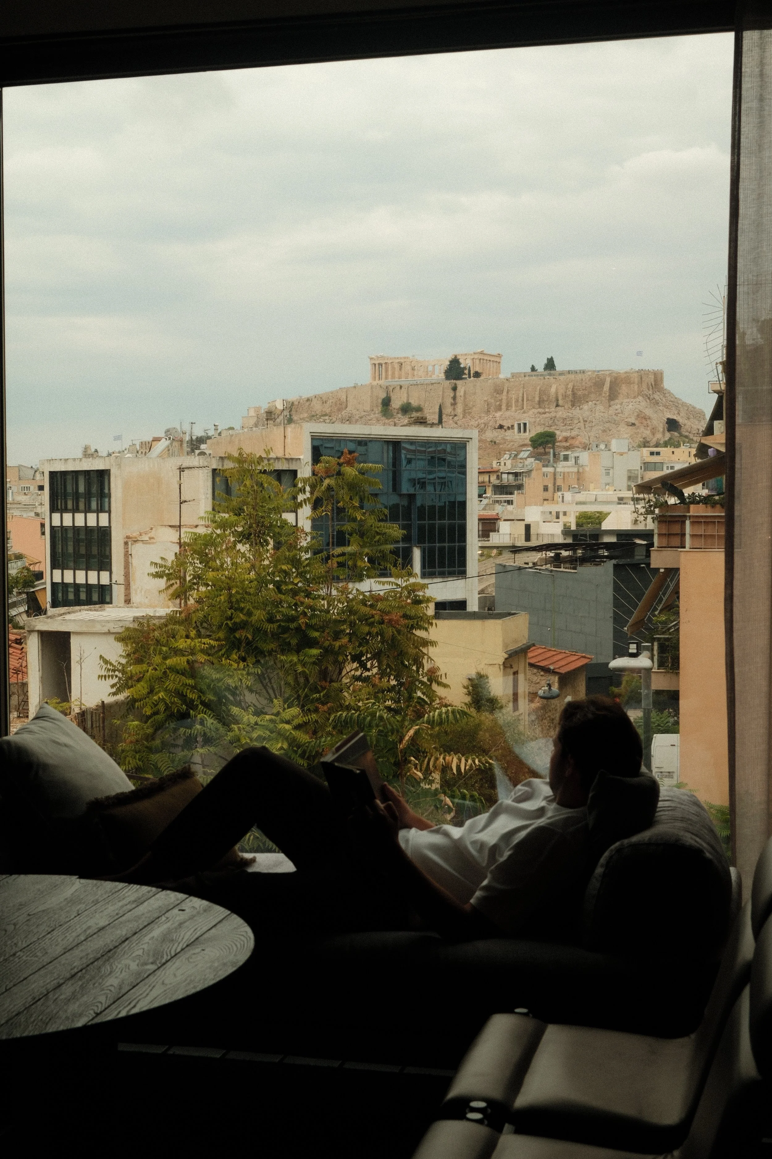 Guest relaxing beside a panoramic window overlooking Athens and the Acropolis