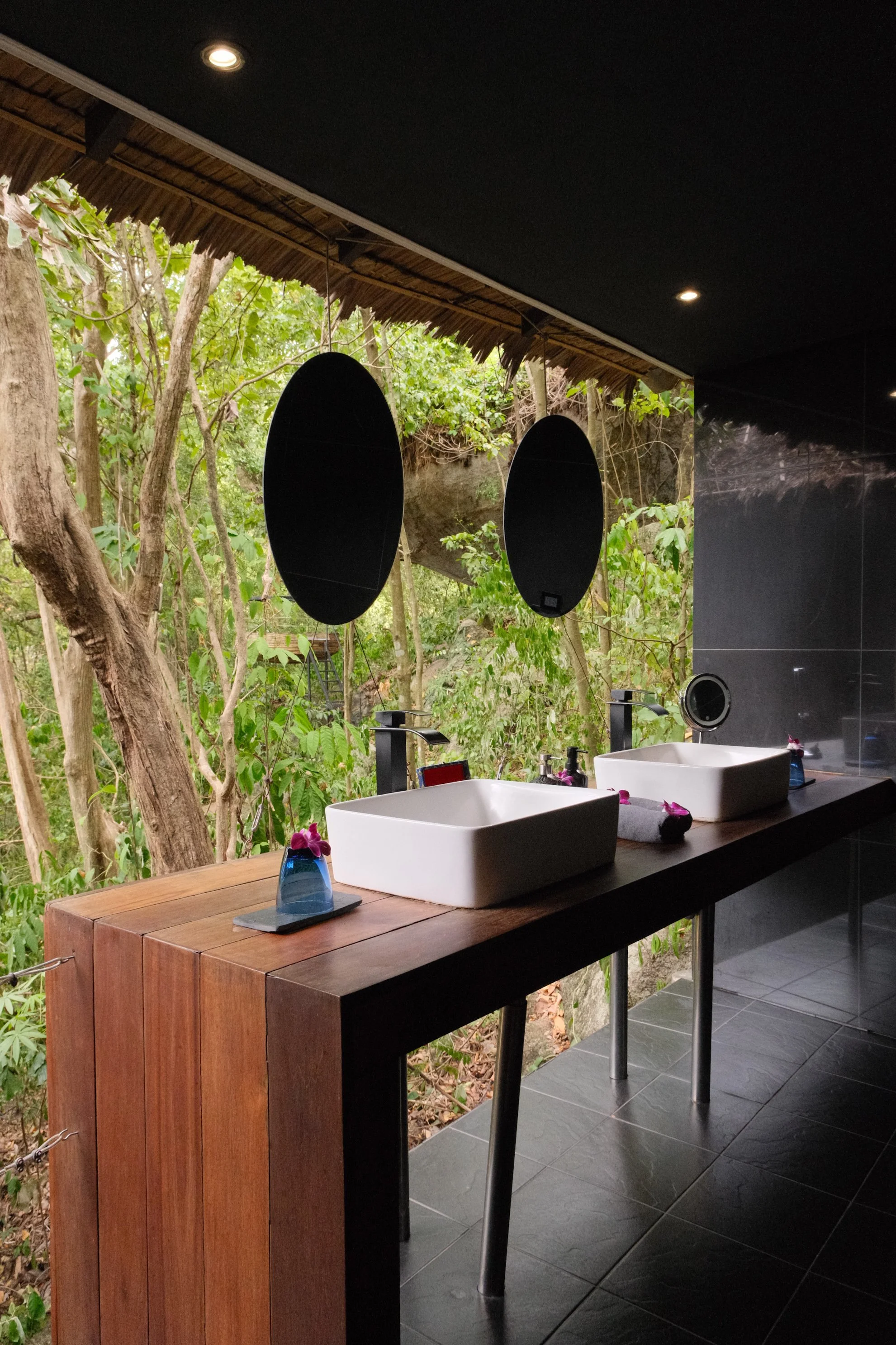 Open-air bathroom with twin vessel sinks, dark finishes, natural wood, and uninterrupted tropical forest views