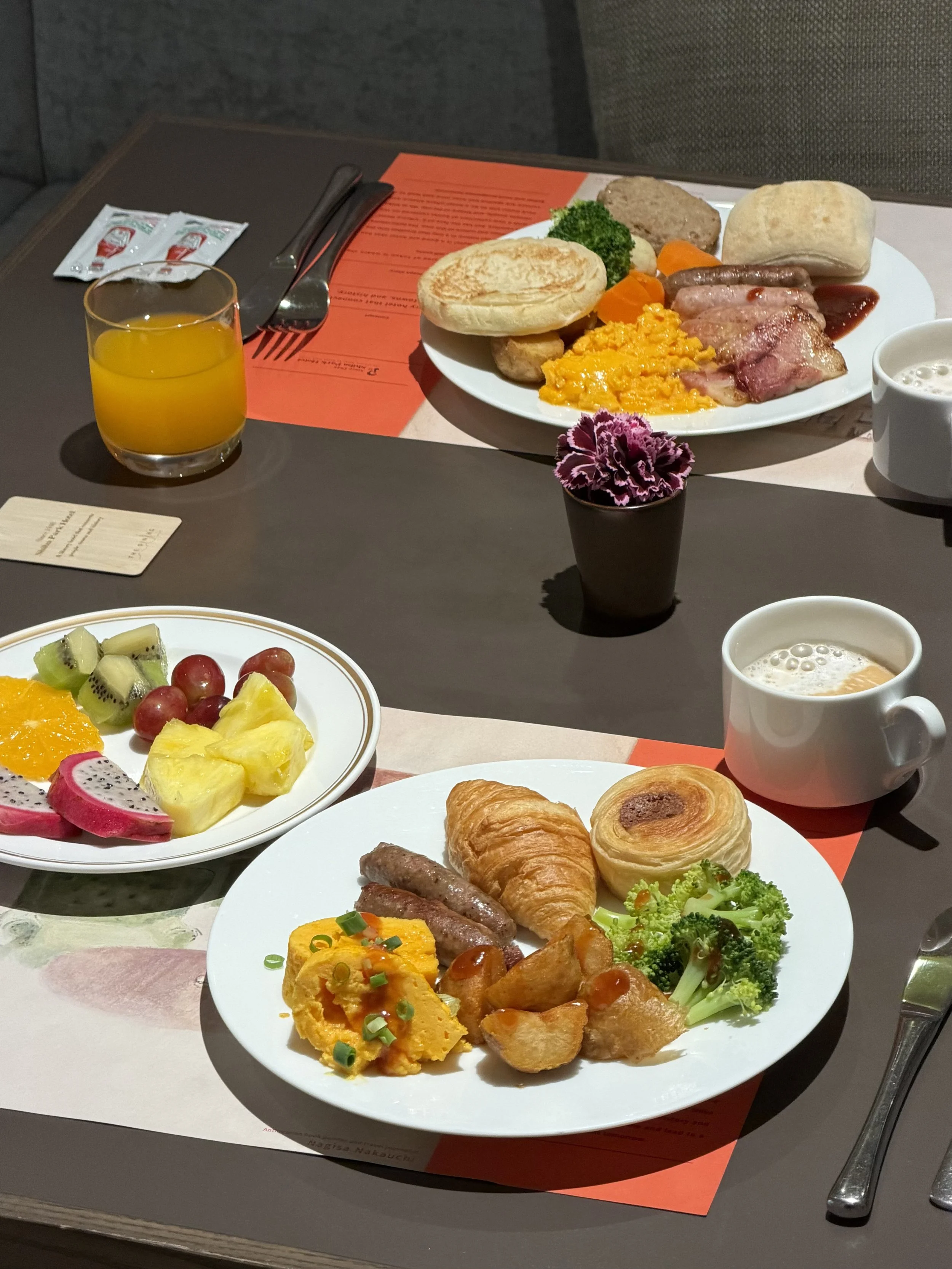 Breakfast spread with pastries, eggs, sausages, fresh fruit, vegetables, coffee, and orange juice.