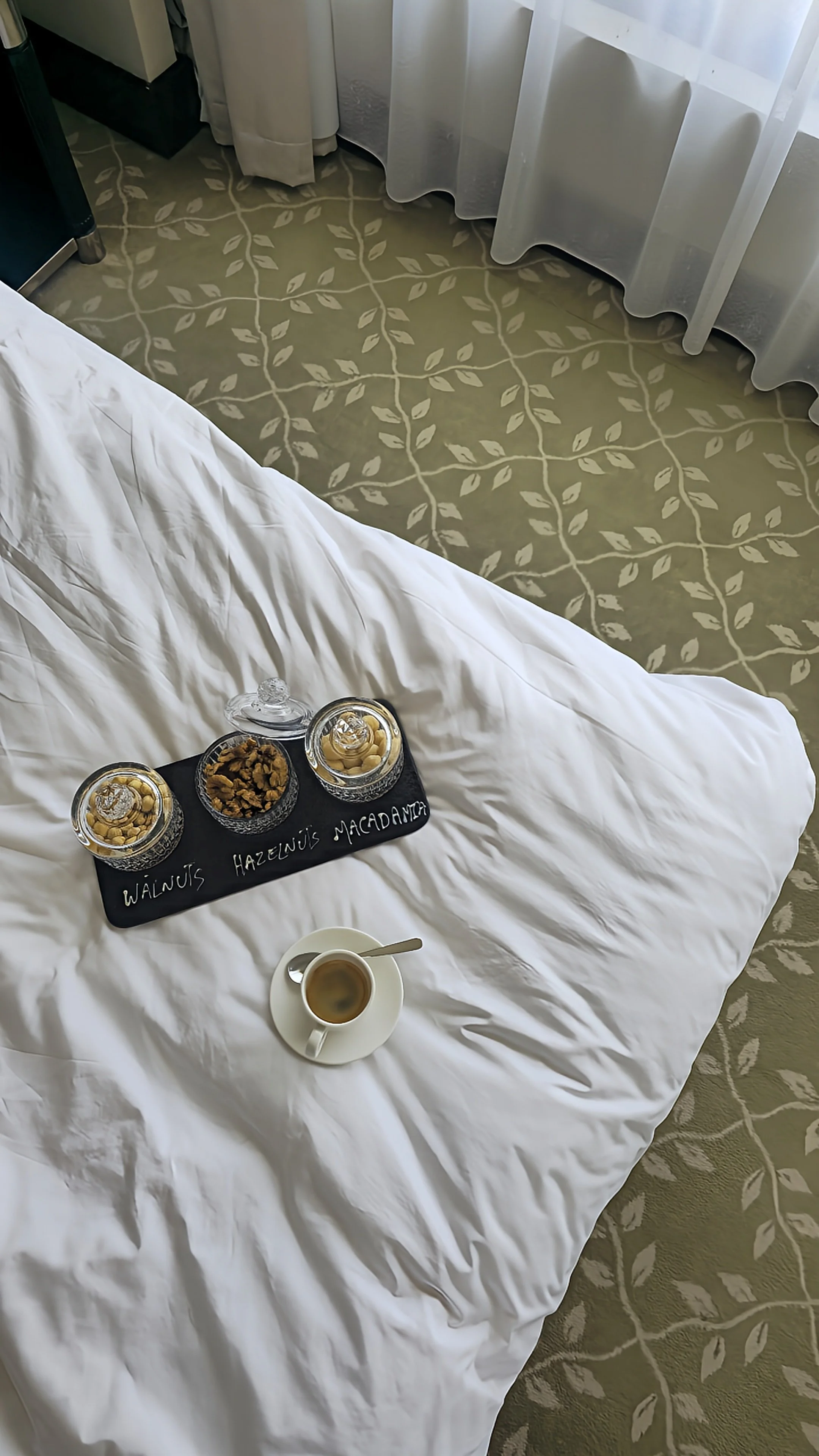 Selection of walnuts, hazelnuts, and macadamia nuts served on a tray beside a cup of coffee on white hotel bedding