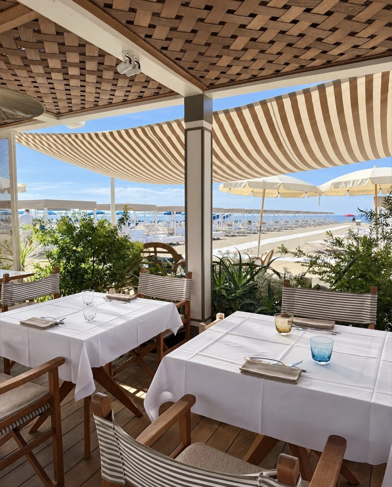 Beachfront restaurant terrace with white tablecloths, striped awnings, and views across the sandy beach