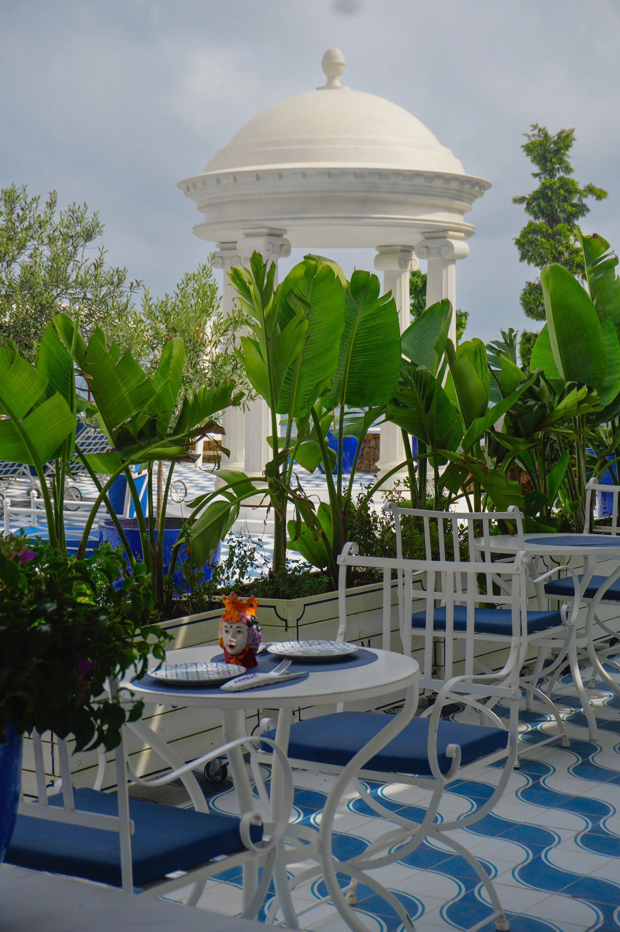 Mediterranean restaurant terrace featuring white wrought iron furniture, blue cushions, tropical greenery, and a decorative pavilion