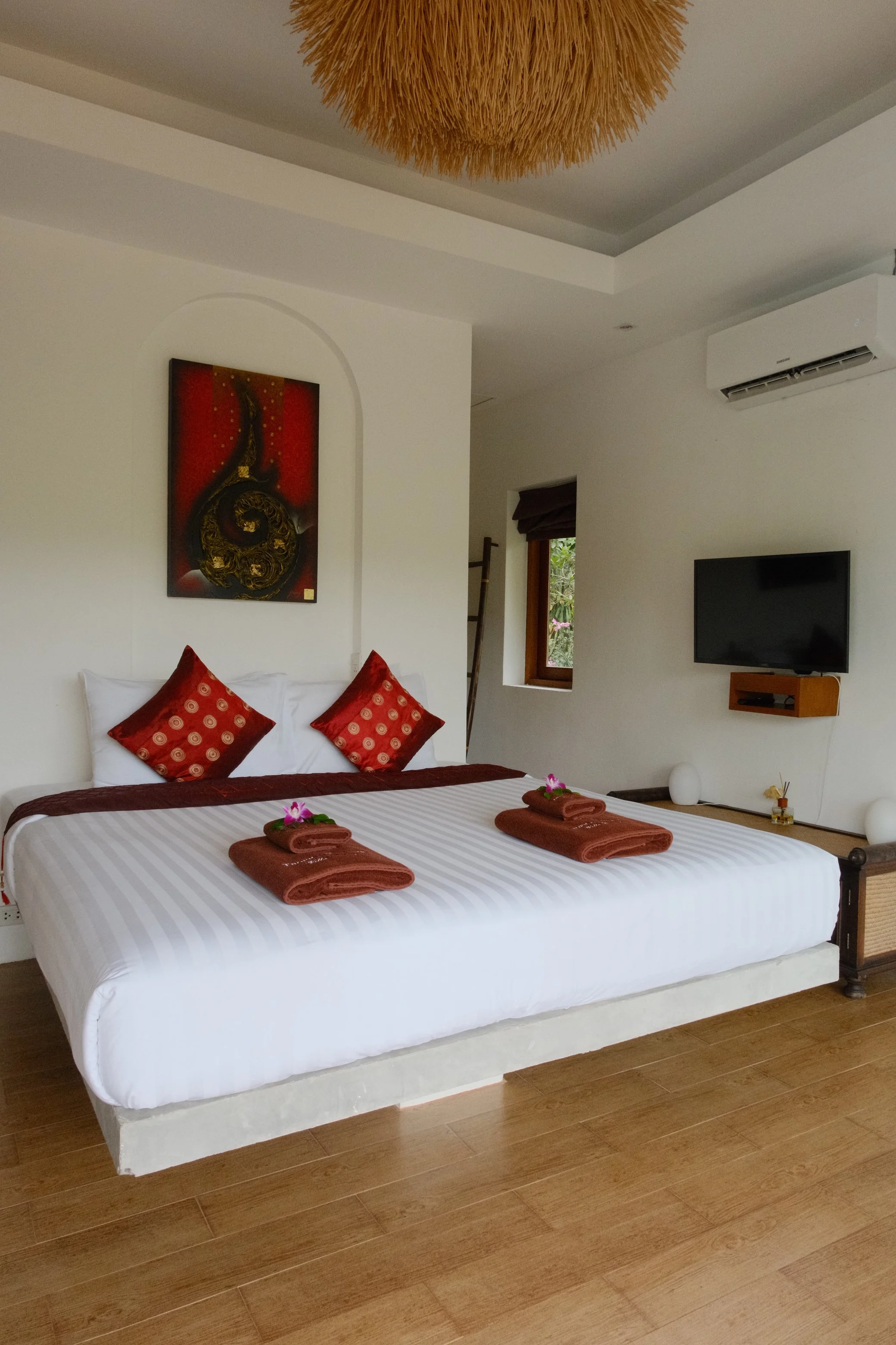 Bright bedroom with red accent cushions, floral details, natural wood flooring, and Thai-inspired artwork