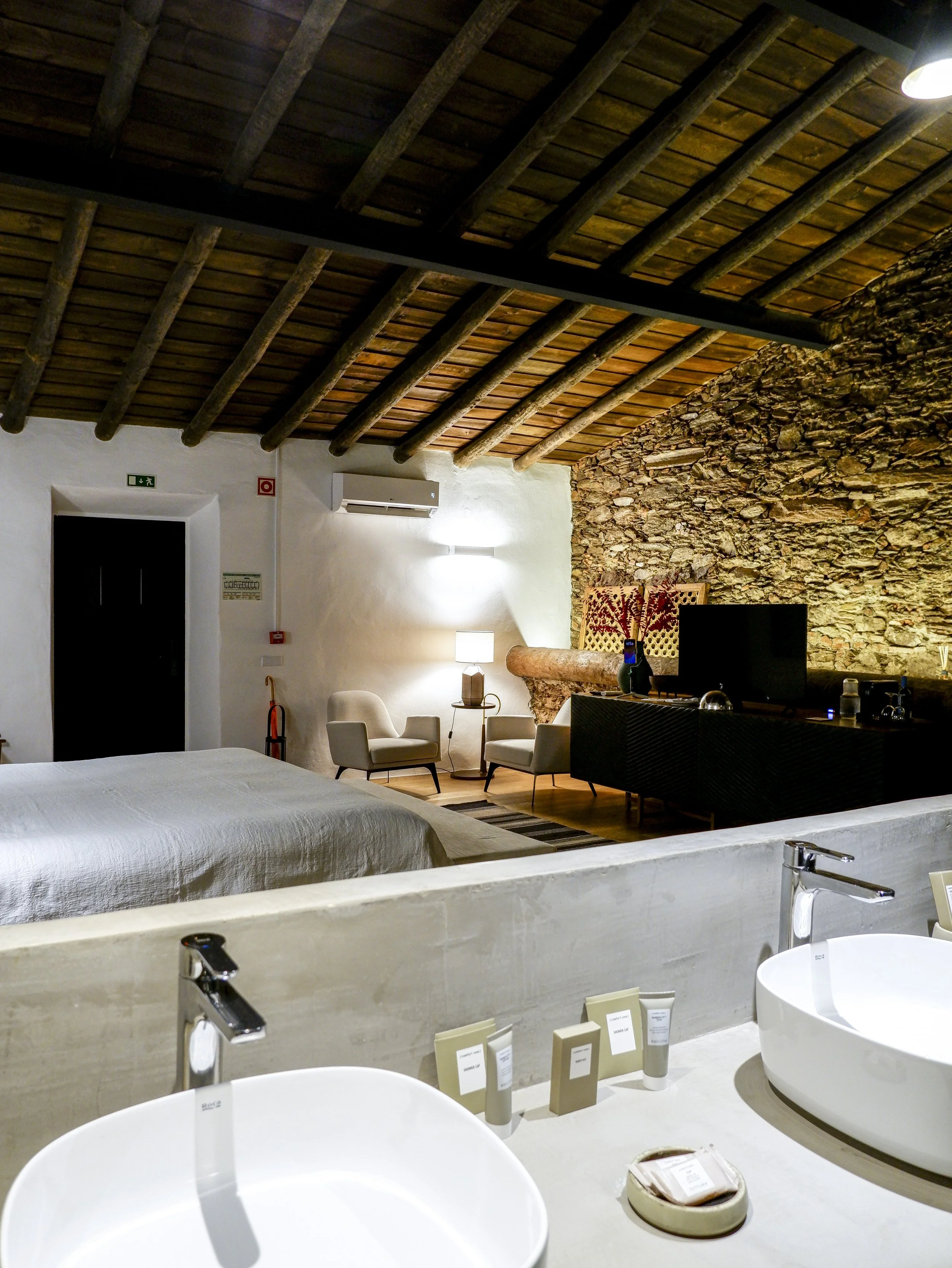 Open-plan bedroom and bathroom with twin stone sinks, natural textures, and traditional wooden ceilings blending heritage architecture with contemporary comfort, Land of Alandroal – Alandroal, Portugal.