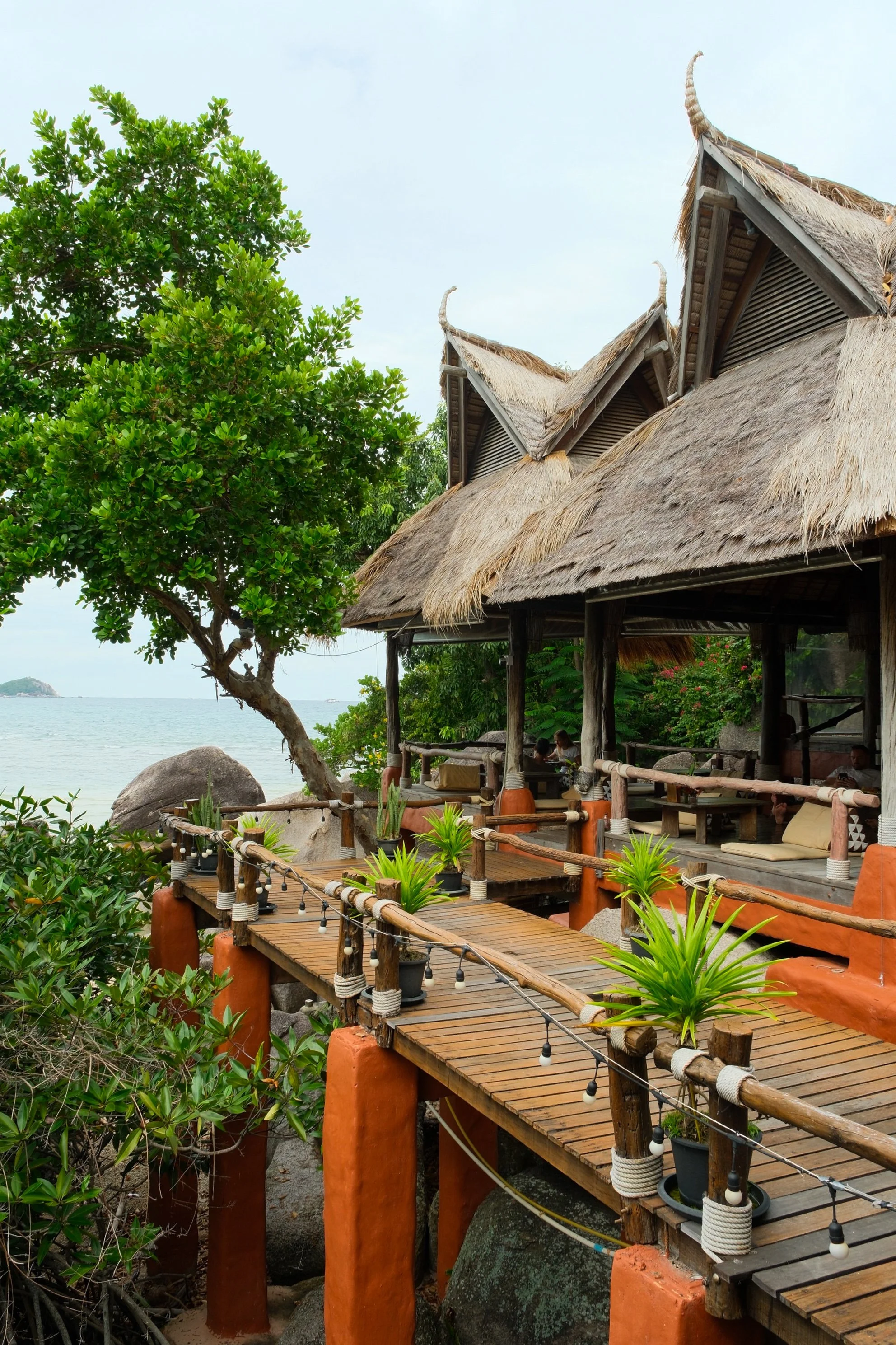 Rustic seaside terrace with thatched roofs, wooden walkways, tropical plants, and ocean views