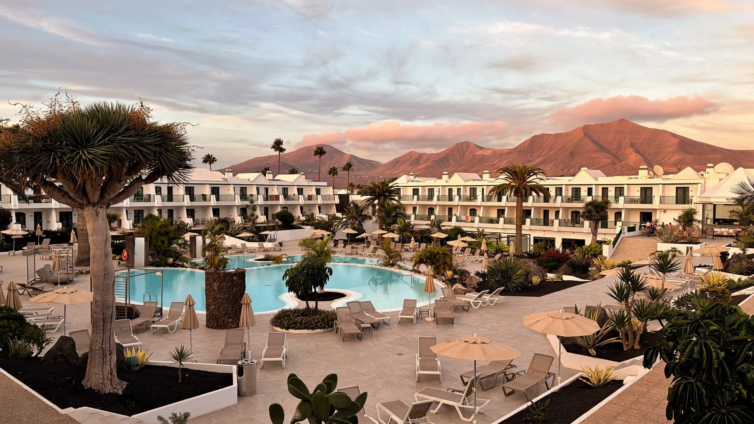 Panoramic view of the resort's swimming pools, sun loungers, and tropical gardens against Lanzarote's volcanic mountains at sunset