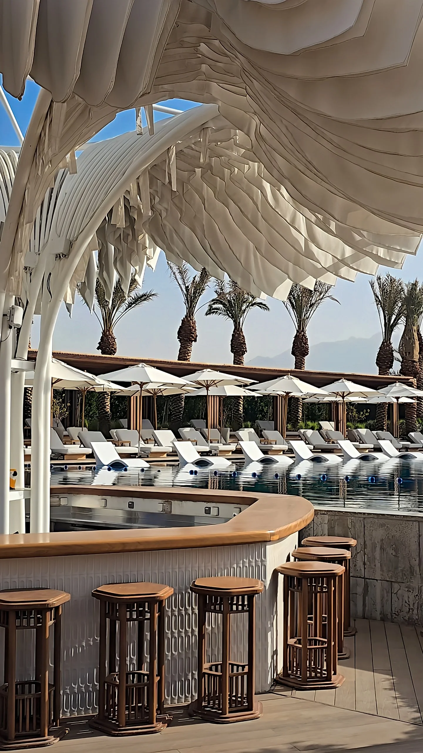 Pool bar with sculptural white canopy, wooden stools, and sun loungers overlooking palm trees and mountains, Hyatt Regency Aqaba Ayla Resort – Aqaba, Jordan.