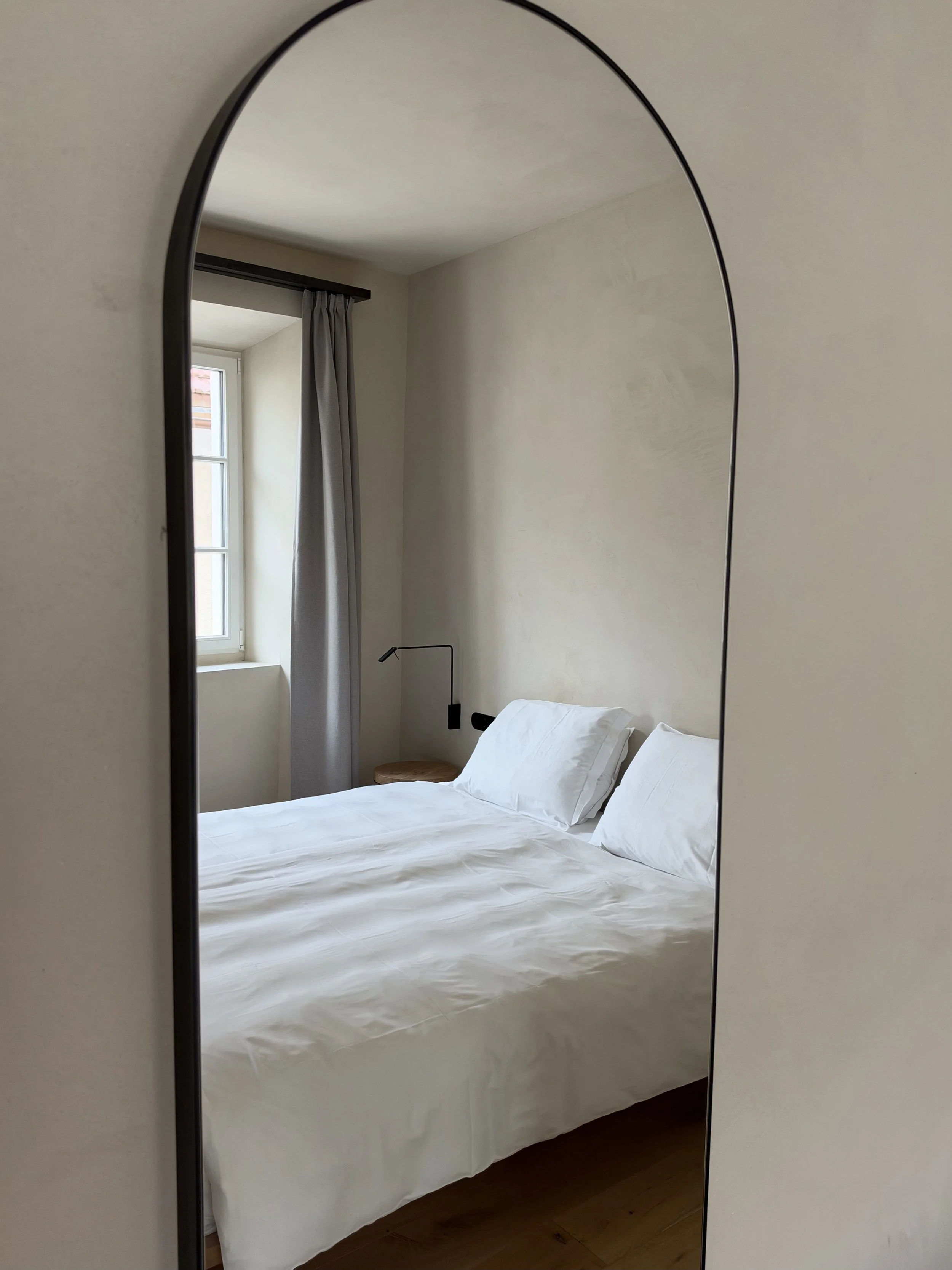 Minimalist guest bedroom reflected in a large arched mirror with white bedding, natural wood floors, and soft lighting