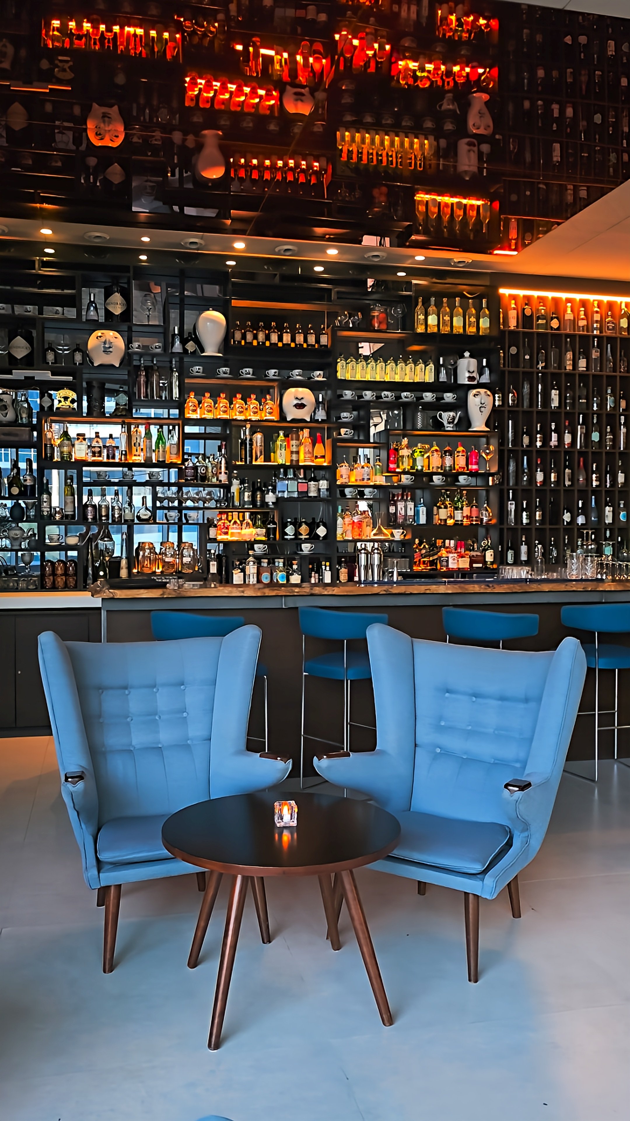Sophisticated hotel bar featuring blue armchairs, a round coffee table, illuminated shelves, and contemporary interior design