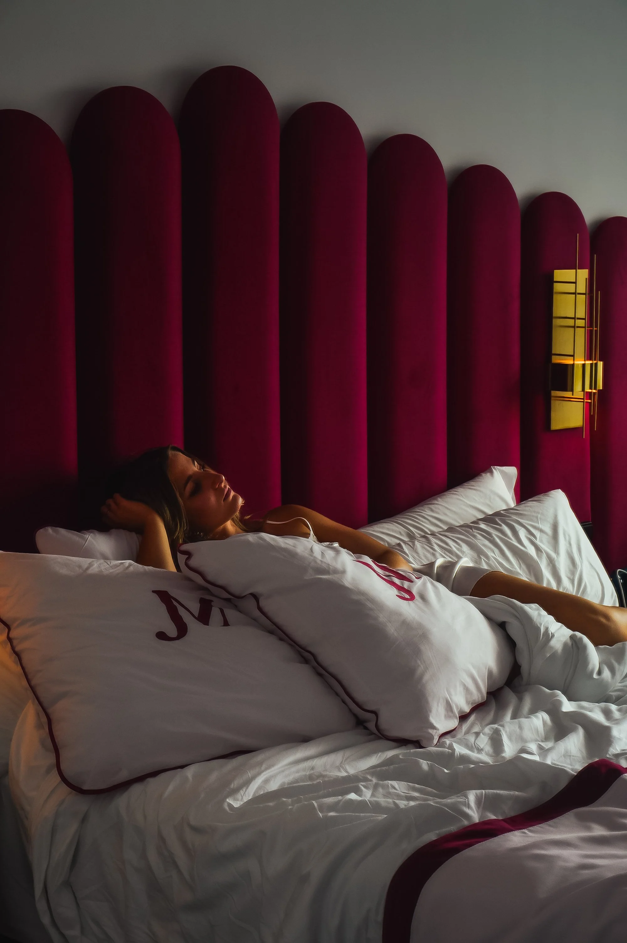 Guest relaxing in a luxurious hotel bed featuring a burgundy velvet headboard, white linens, and elegant golden wall lighting