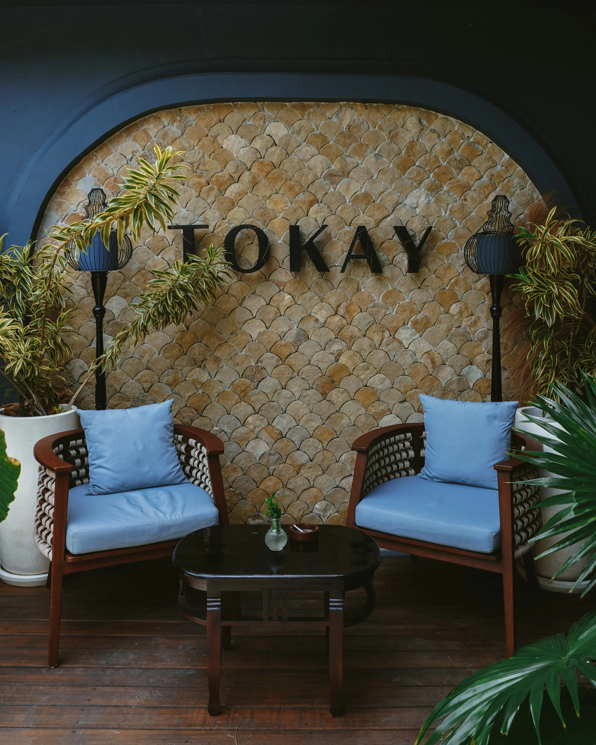 Tropical lounge with woven wood chairs and soft blue cushions beneath a stone-textured wall at Villa Tokay, Gili Air