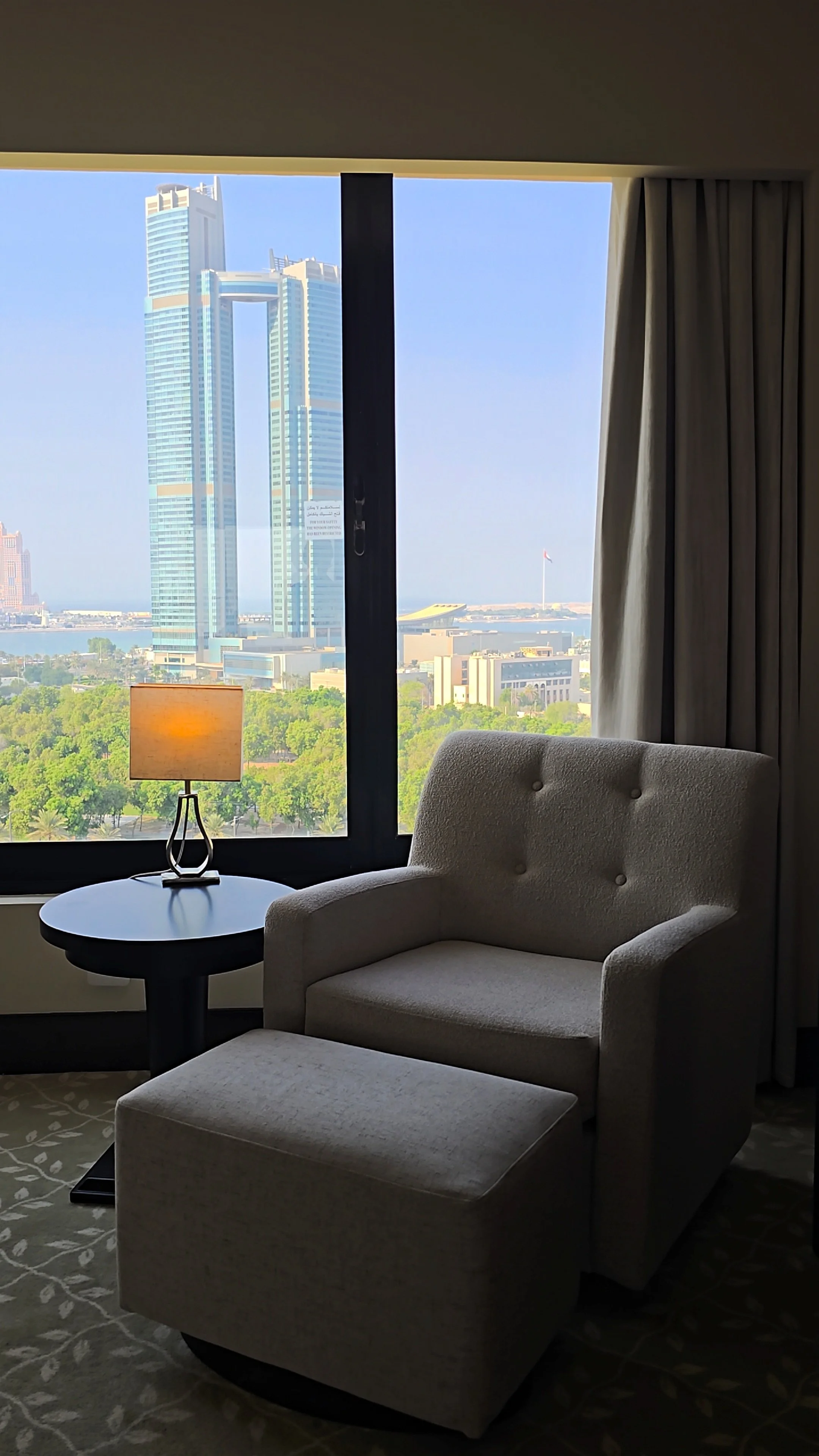 Comfortable guest room seating area with an upholstered armchair, matching ottoman, and panoramic Abu Dhabi skyline views