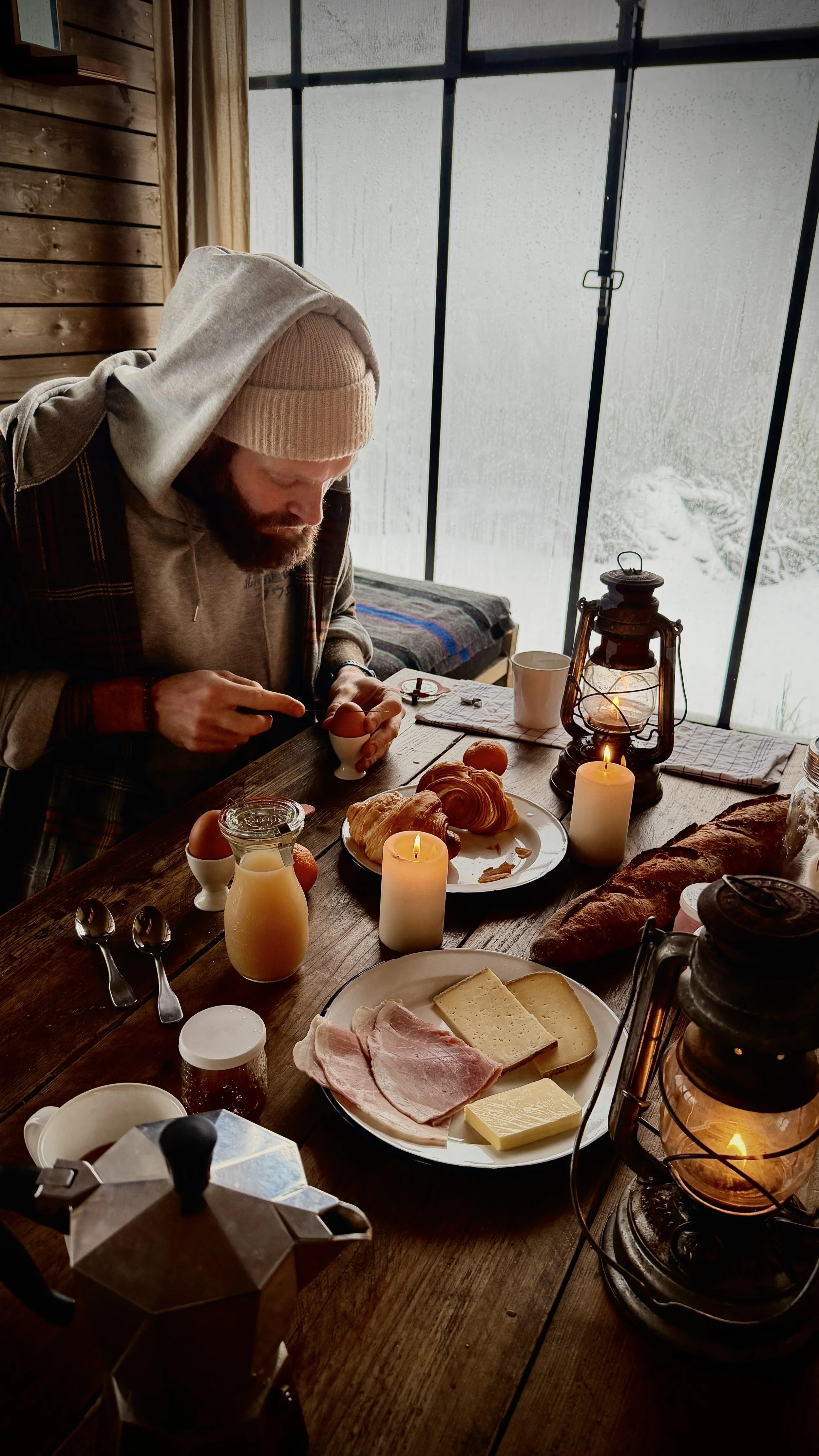 A cozy wooden interior where a guest prepares breakfast by the window as snow falls softly outside, Nutchel Forest Village Alsace – Plaine, France.