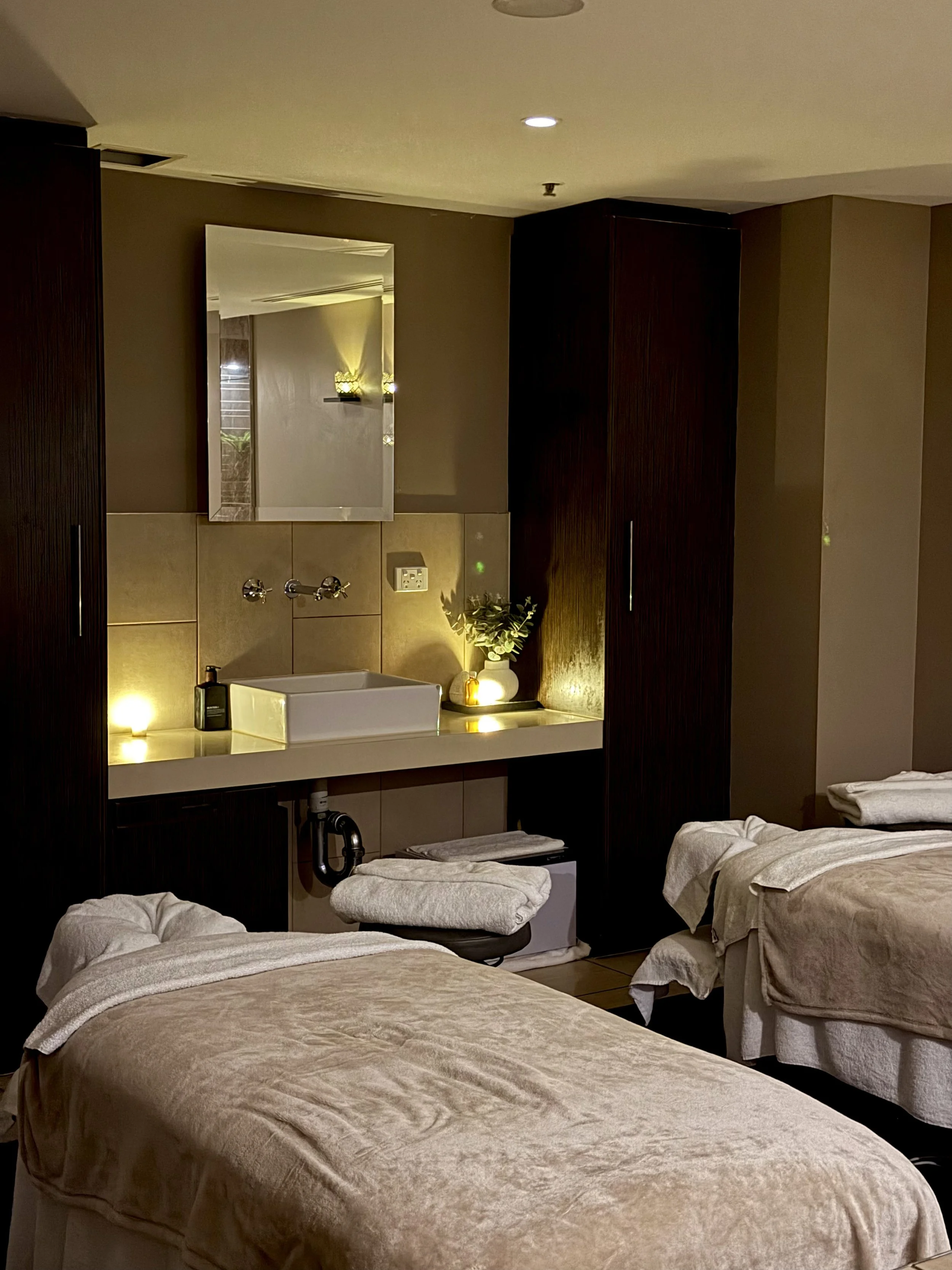 Relaxing spa treatment room featuring two massage beds, soft lighting, dark wooden cabinetry, and a modern vanity