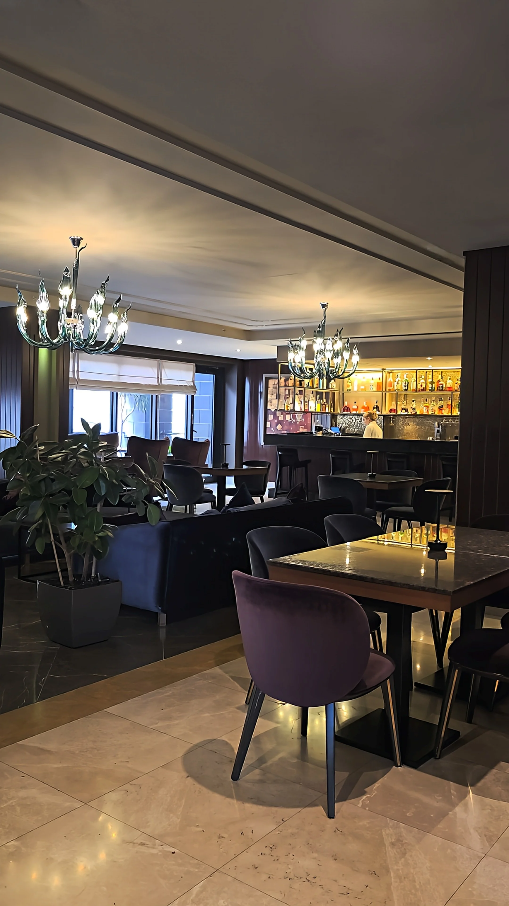 Elegant hotel bar and lounge with contemporary seating, chandeliers, and a backlit drinks display