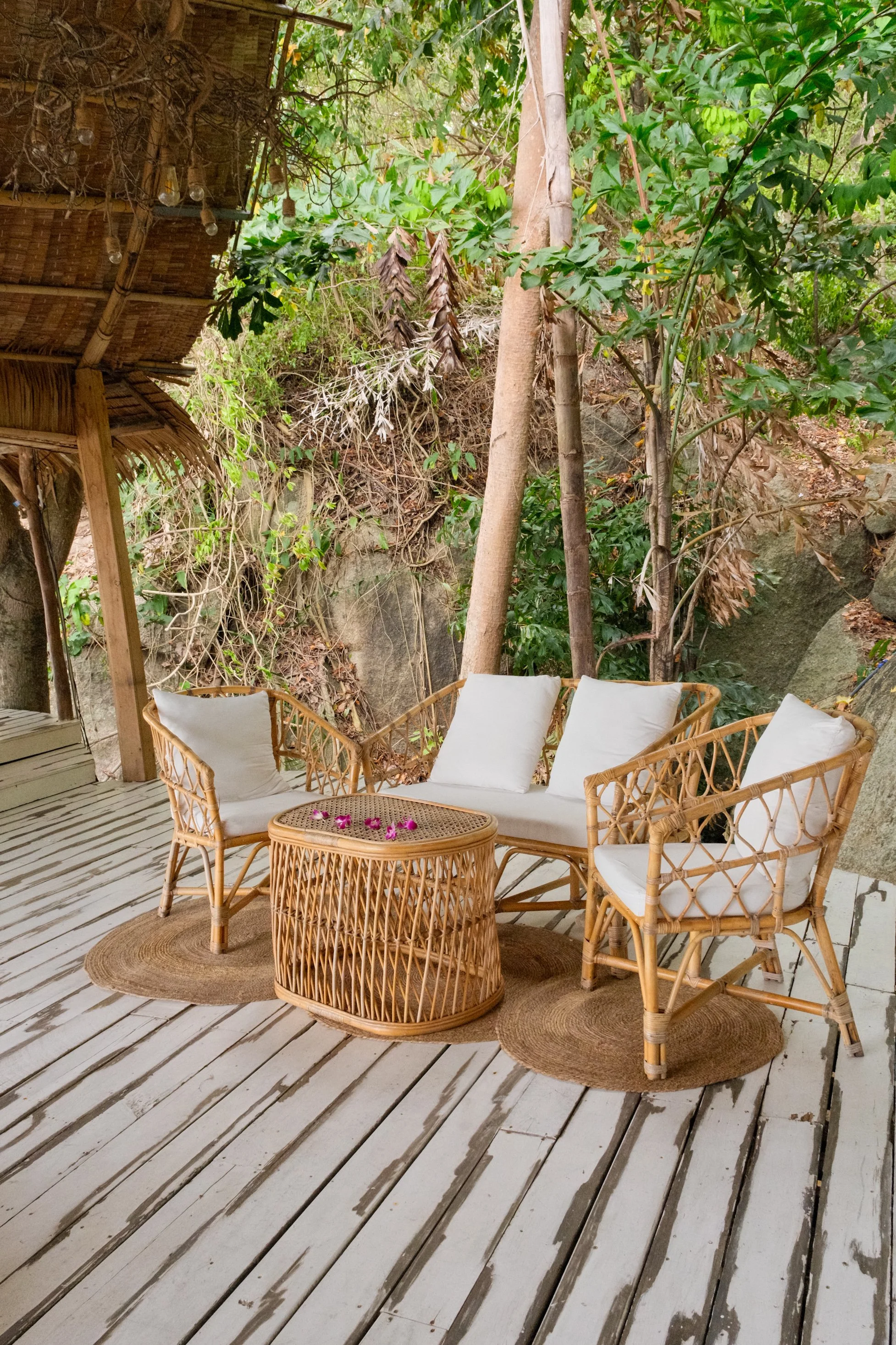 Rattan lounge chairs and woven table arranged on a covered wooden terrace surrounded by dense jungle