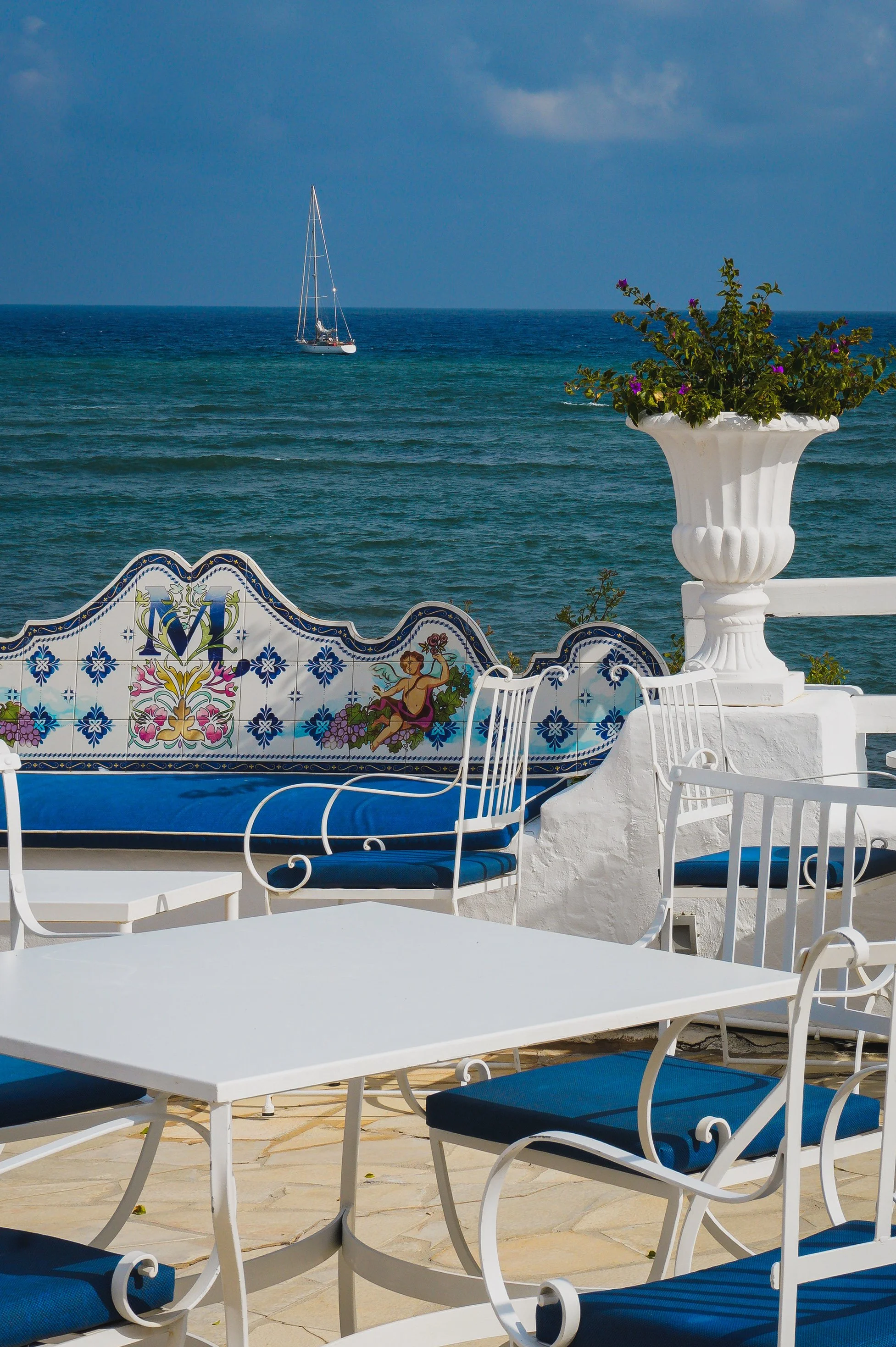 Seafront terrace with blue and white seating, decorative ceramic tiles, and a sailboat overlooking the Mediterranean Sea