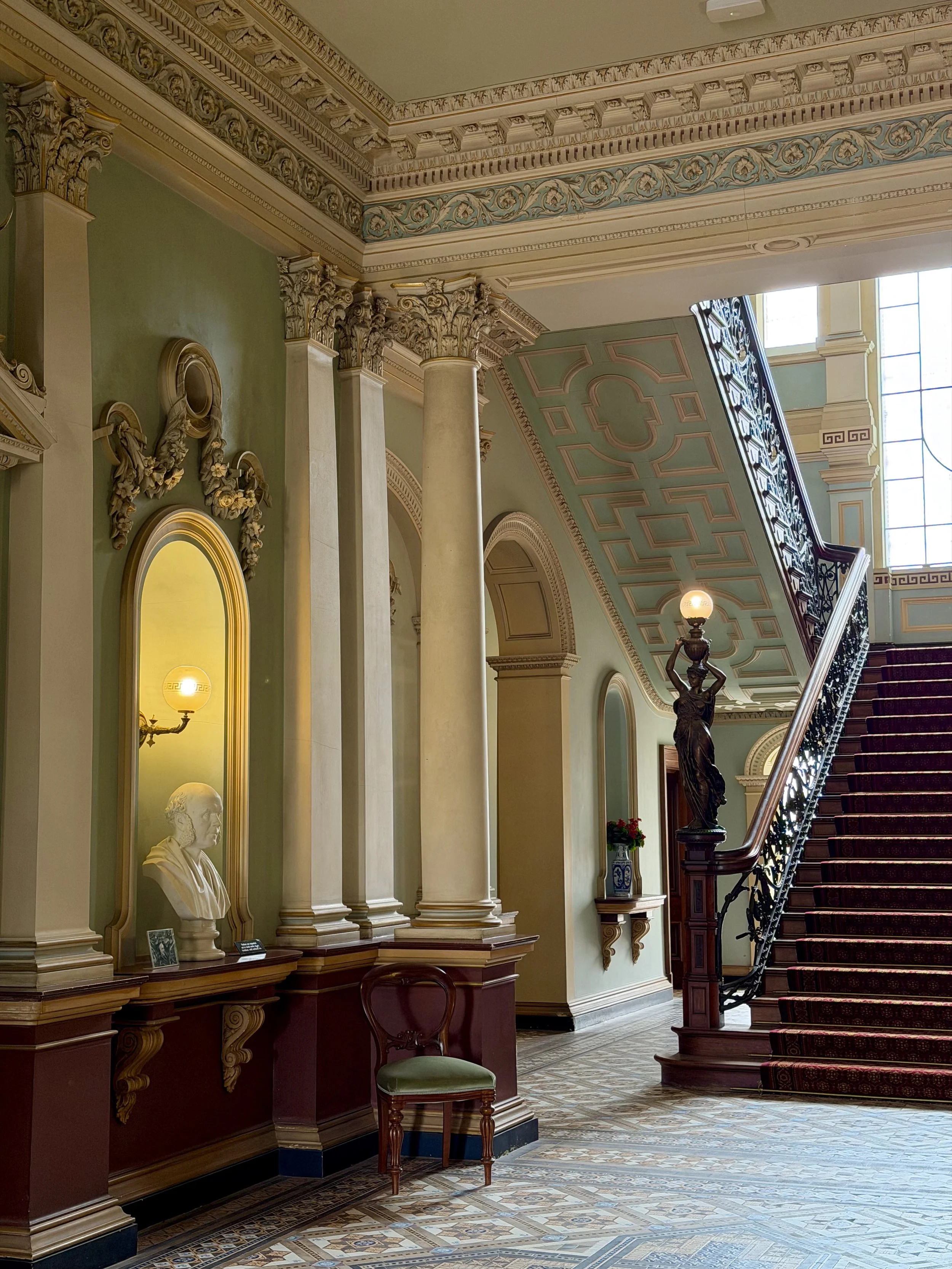 Elegant heritage staircase featuring ornate columns, decorative ceilings, patterned flooring, and intricate iron railings