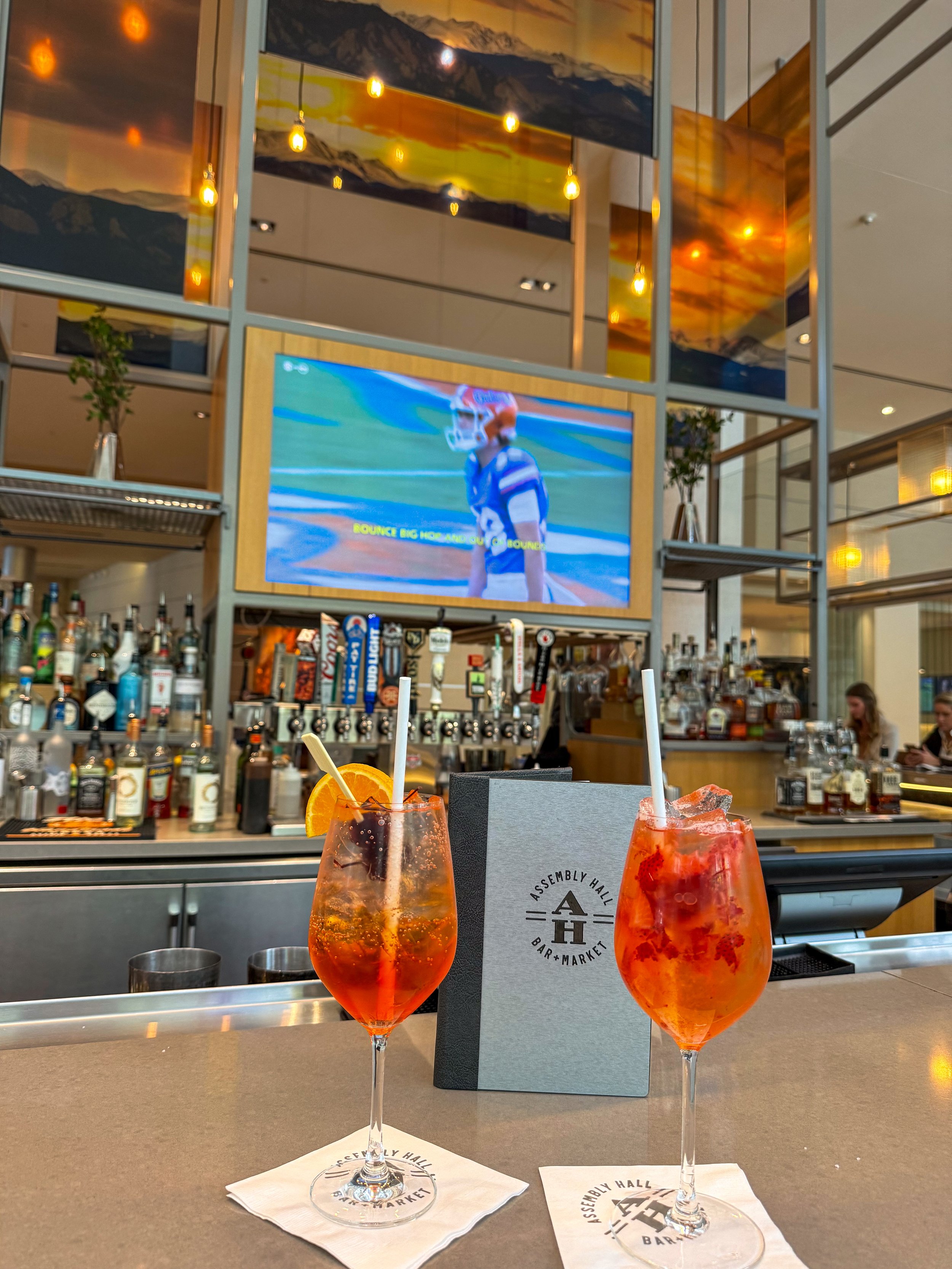 Two colorful cocktails served at Assembly Hall Bar + Market with a modern bar and large television in the background