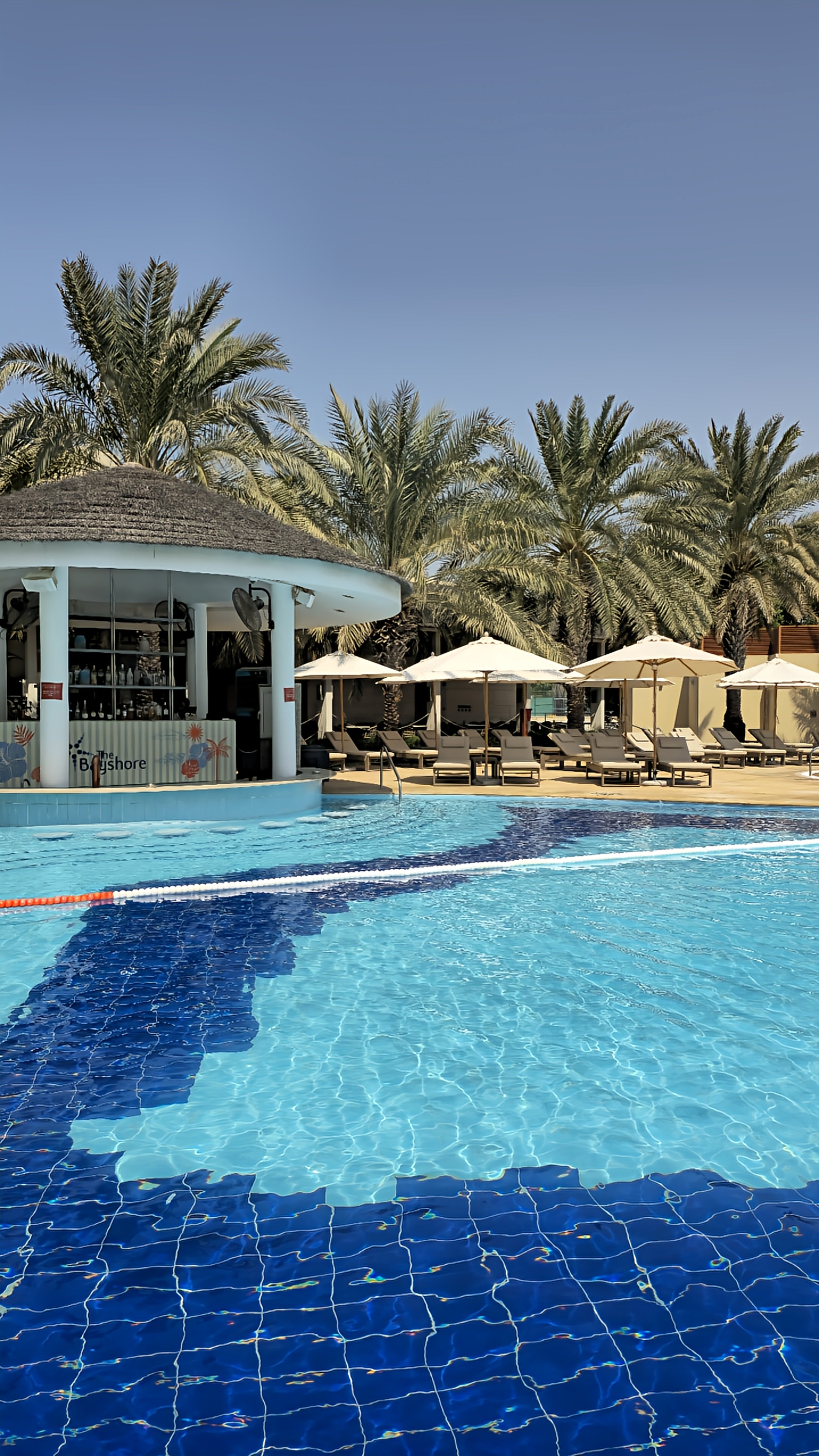 Poolside bar surrounded by tropical palm trees, sun loungers, white umbrellas, and crystal-clear swimming pool water