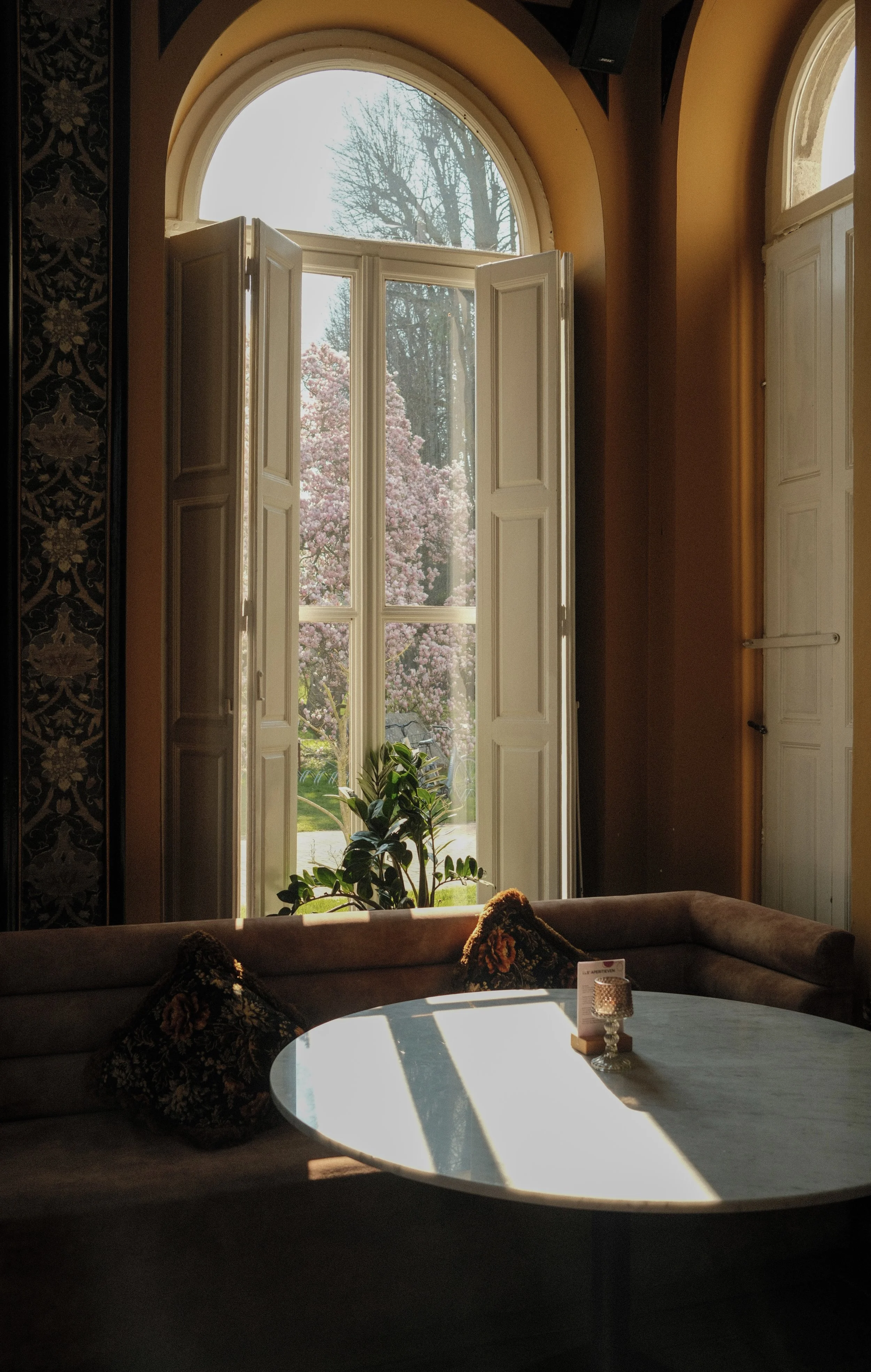 Sunlit lounge with arched windows, soft seating, and garden views creating a calm and inviting atmosphere, Château St. Gerlach – Vaeshartelt Maastricht.
