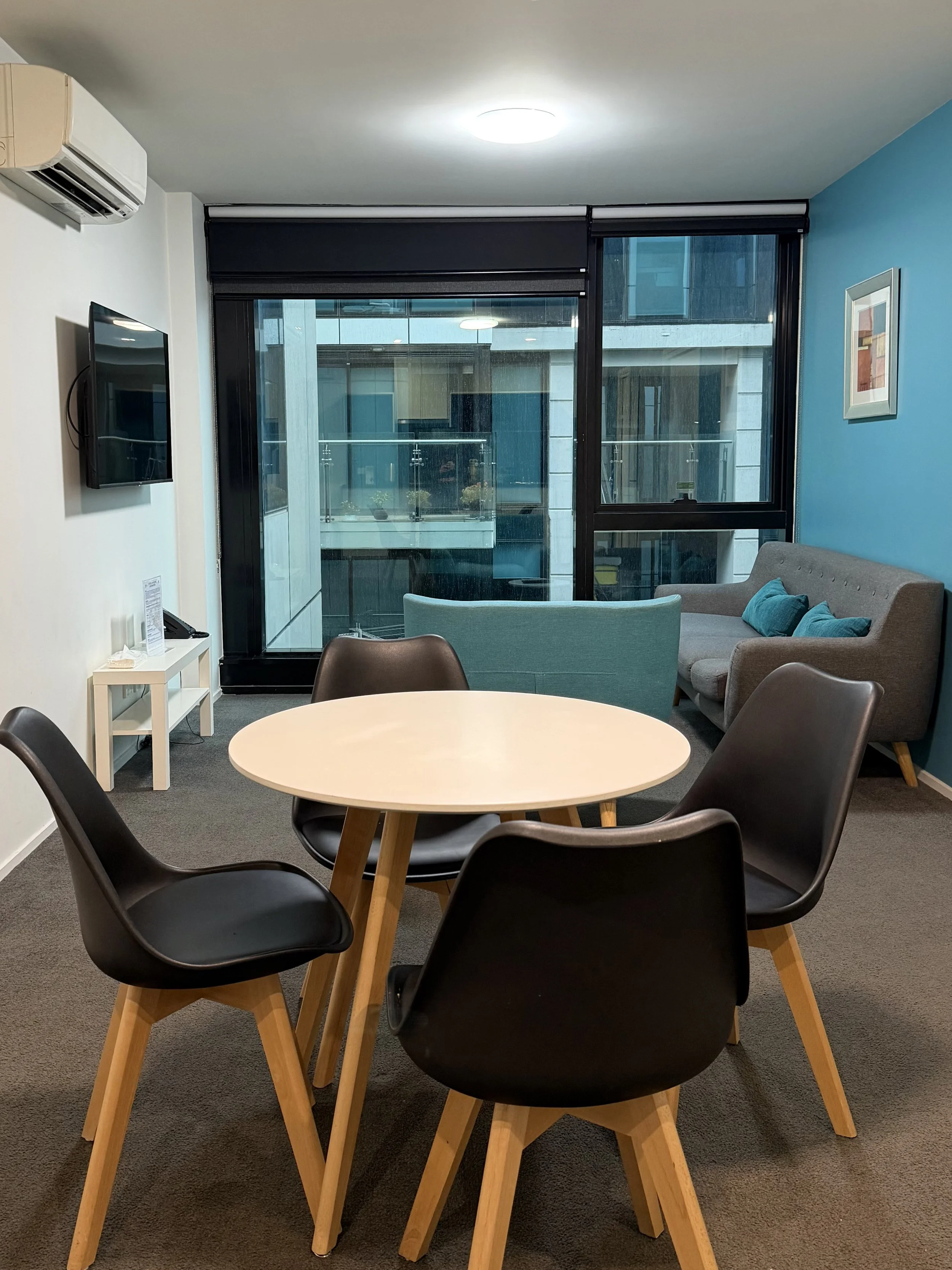 Spacious apartment living area featuring a round dining table, grey sofa, blue accent wall, and floor-to-ceiling windows