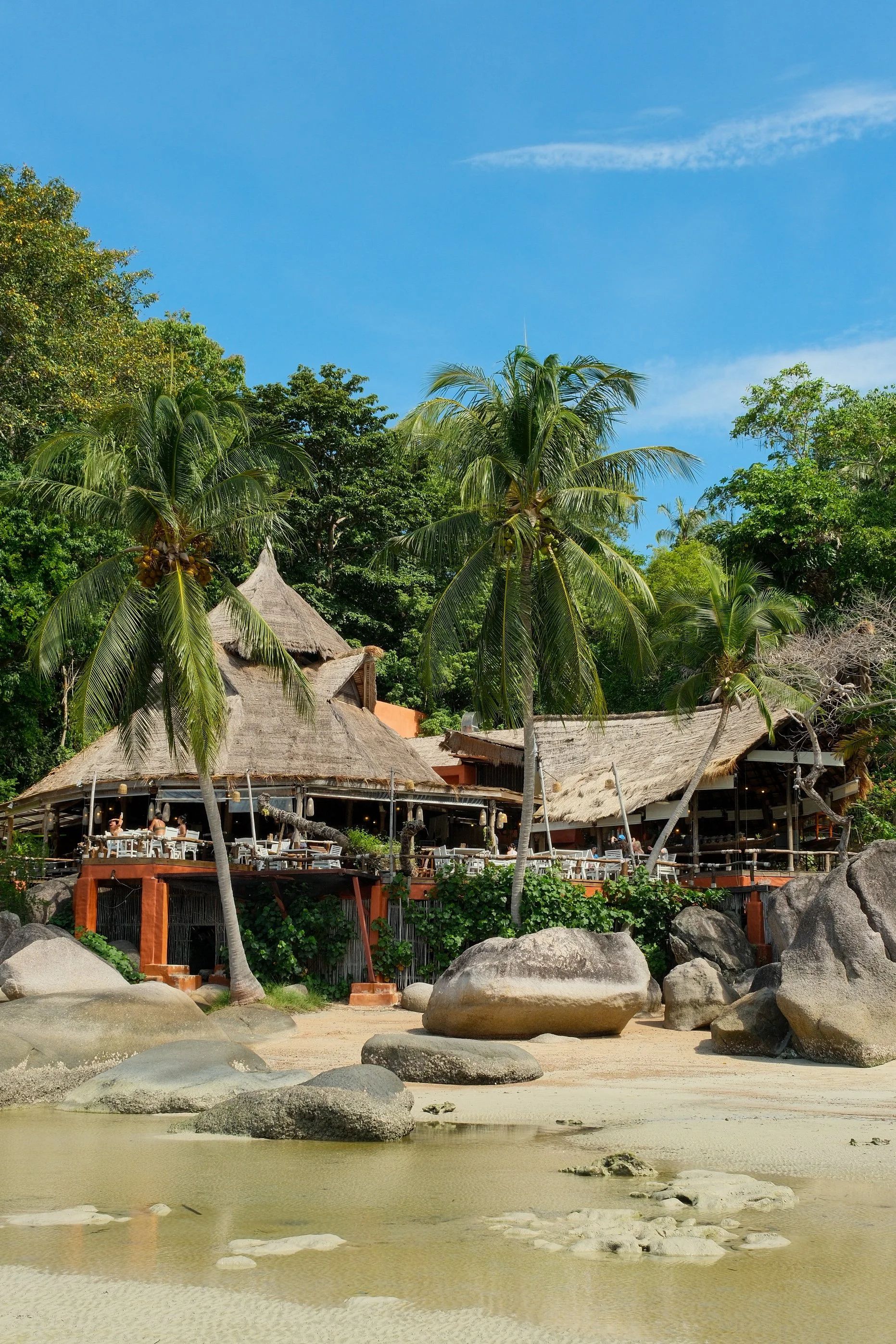 Beachfront restaurant framed by coconut palms, granite boulders, and traditional thatched roofs