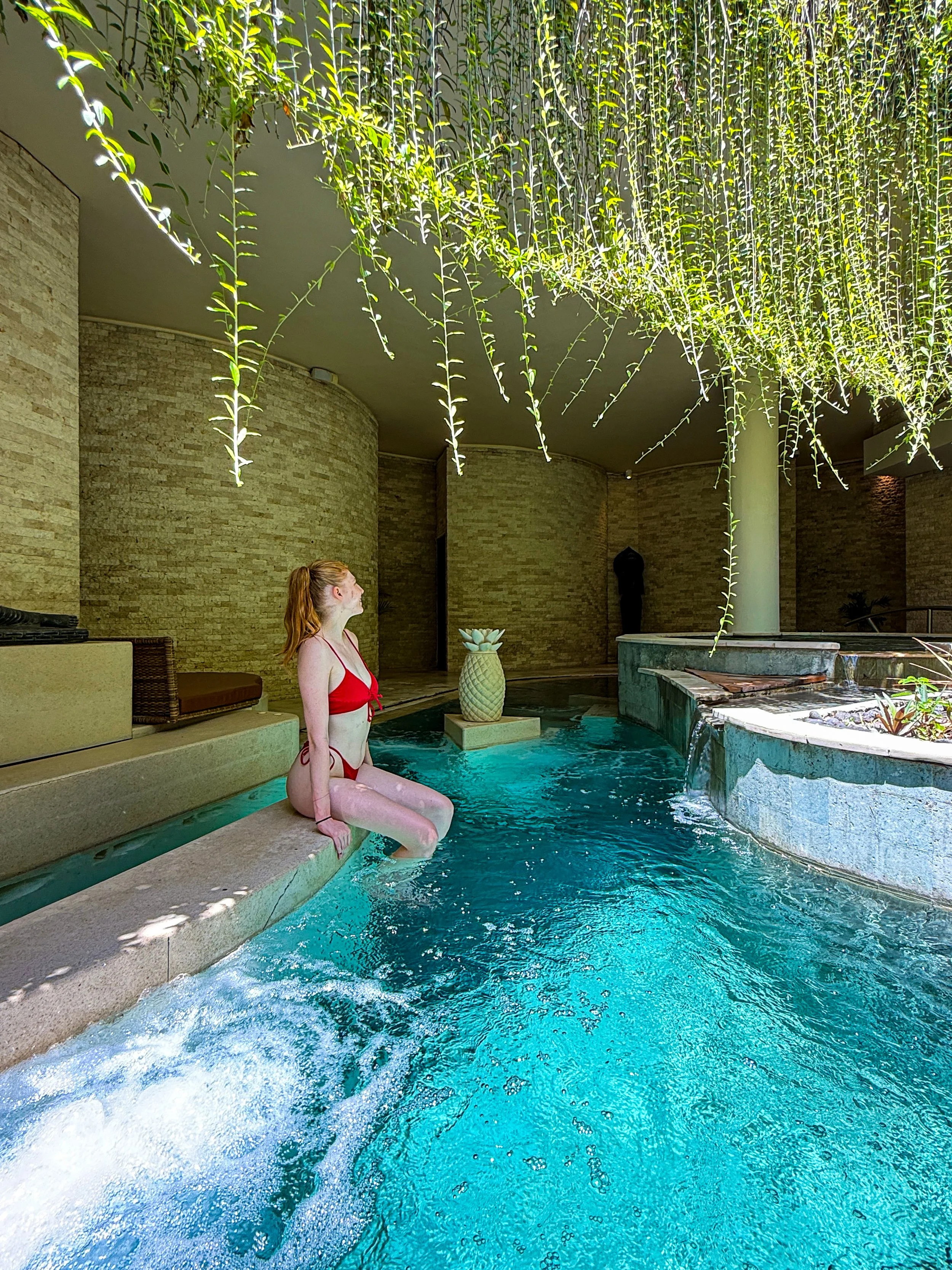 An indoor wellness pool surrounded by cascading water features creates a relaxing spa experience beneath hanging greenery